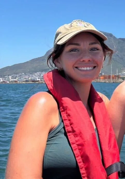 Woman smiling with a cap and life vest, near water with mountains in the background.