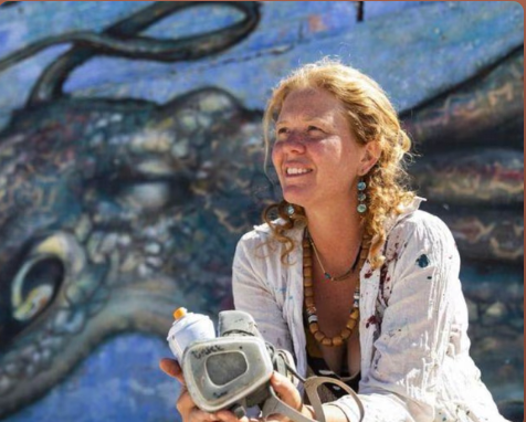 A woman with curly blonde hair and earrings, wearing a white shirt, sits in front of a colorful mural of a dragon, holding a camera.