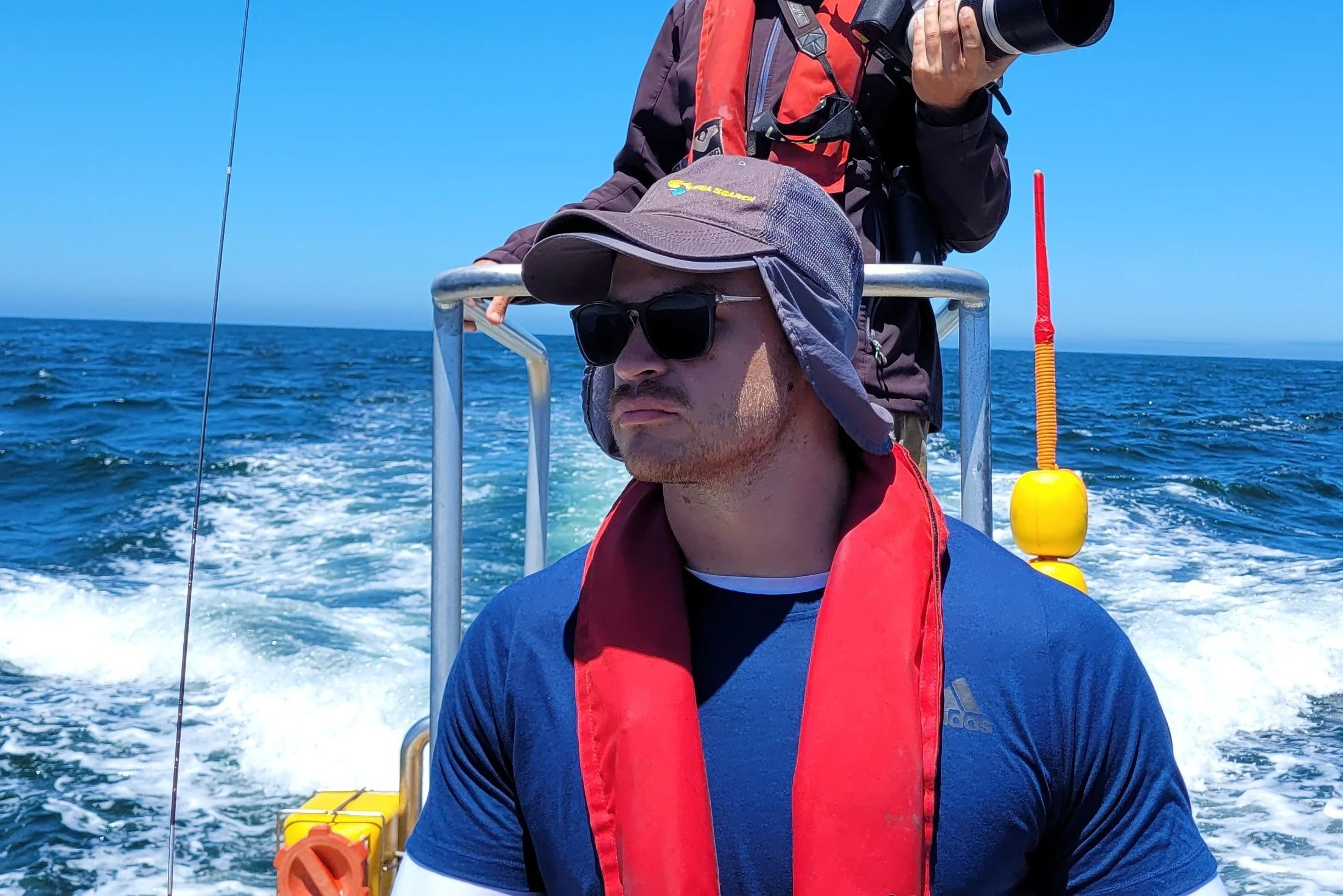 Two men on a boat, one with sunglasses and a hat, the other with a camera, sailing on the open ocean on a sunny day.