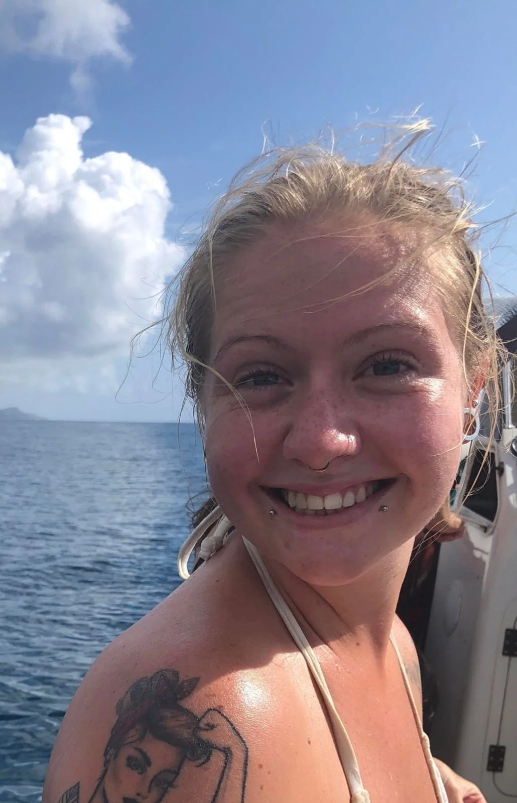 A smiling young woman with blonde hair, blue eyes, and piercings on her lips and nose, on a boat in the ocean on a partly cloudy day.