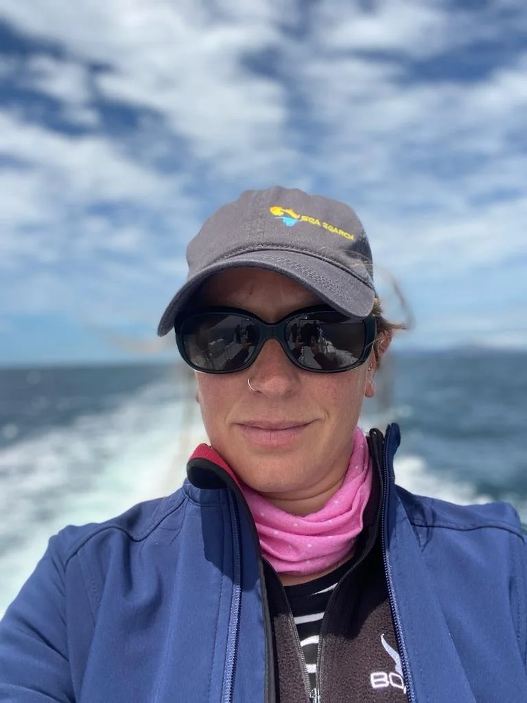Woman wearing sunglasses, a baseball cap, and a pink neck gaiter, taking a selfie on a boat with ocean and cloudy sky in the background.