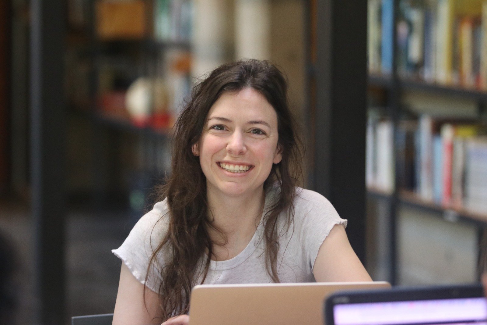 A young woman with long brown hair smiling at the camera, sitting in a library with shelves of books in the background.