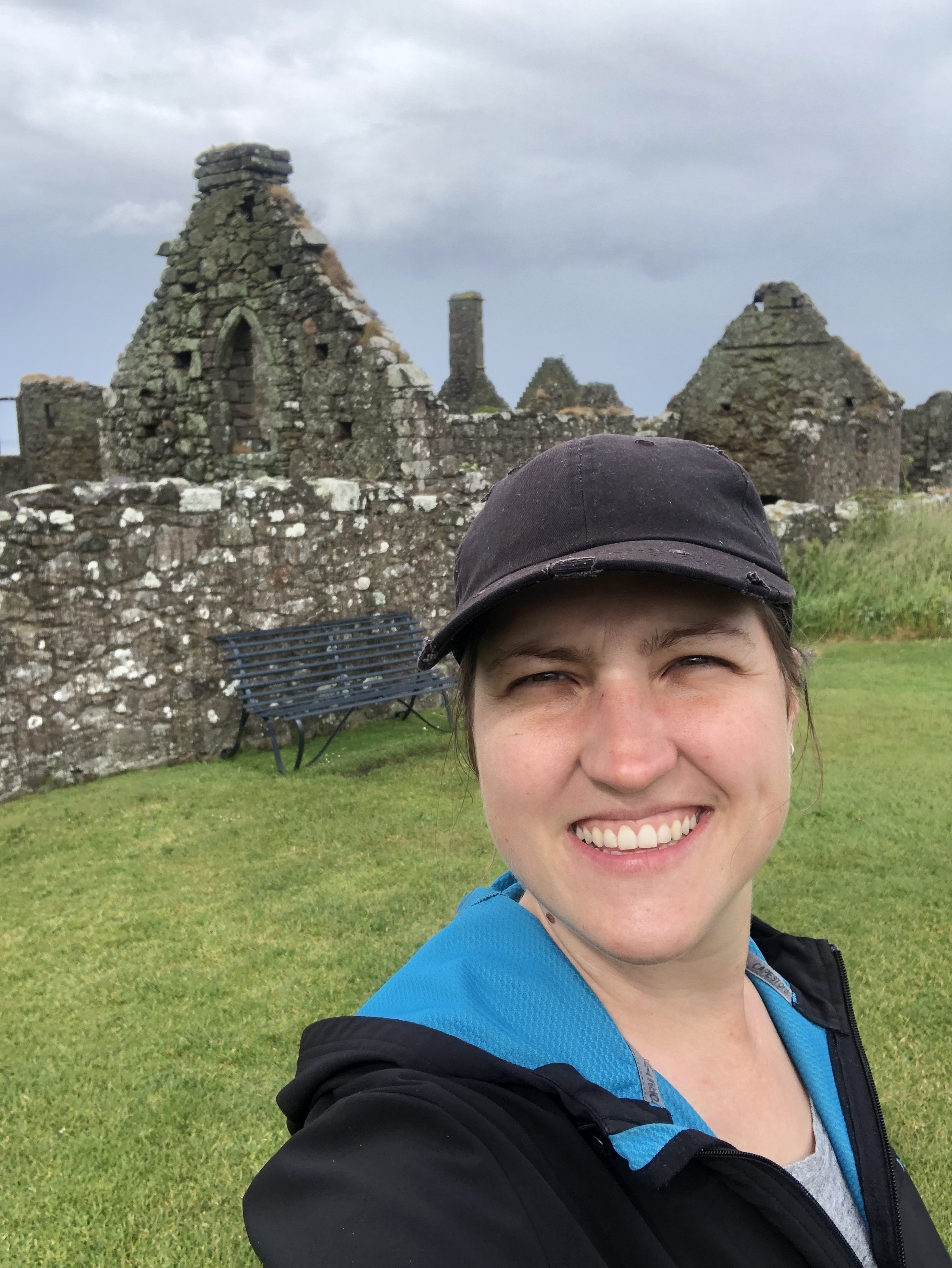 Smiling woman wearing a black and blue jacket and a black cap, taking a selfie in front of ancient stone ruins on cloudy day with grass and a black metal bench nearby.