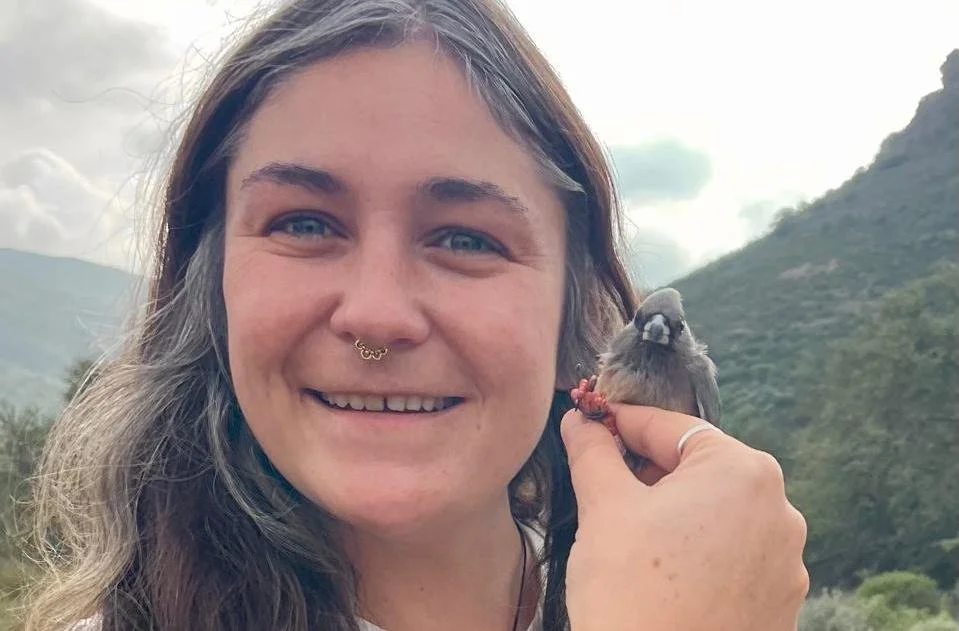 A woman with long hair holding a small bird close to her face outdoors with mountains and cloudy sky in the background.