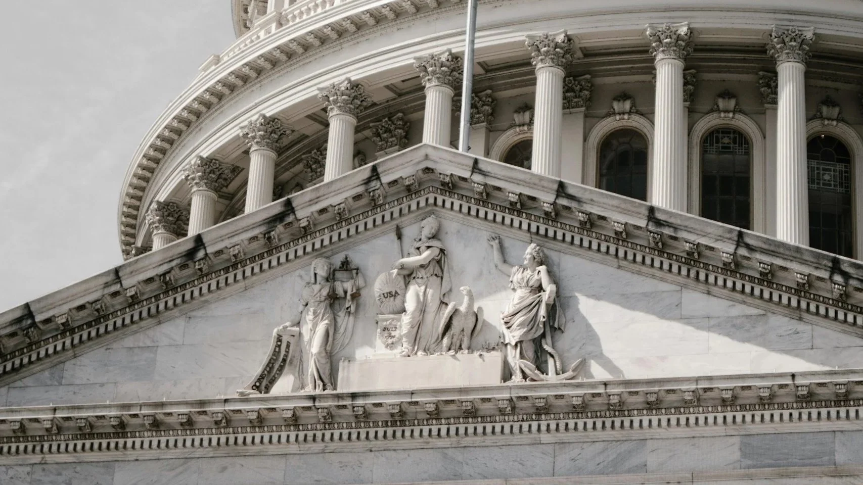 Close-up of the architectural details on the United States Capitol building, featuring a frieze sculpture with draped female figures and a bald eagle, surrounded by classical columns and ornate carvings.