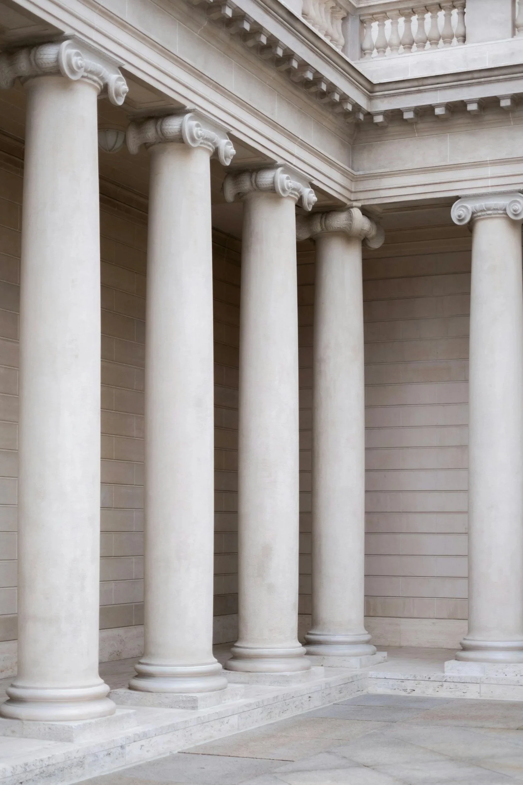View of white marble columns with Ionic capitals in a classical building, featuring ornate detailing and a balustrade balcony at the upper level.
