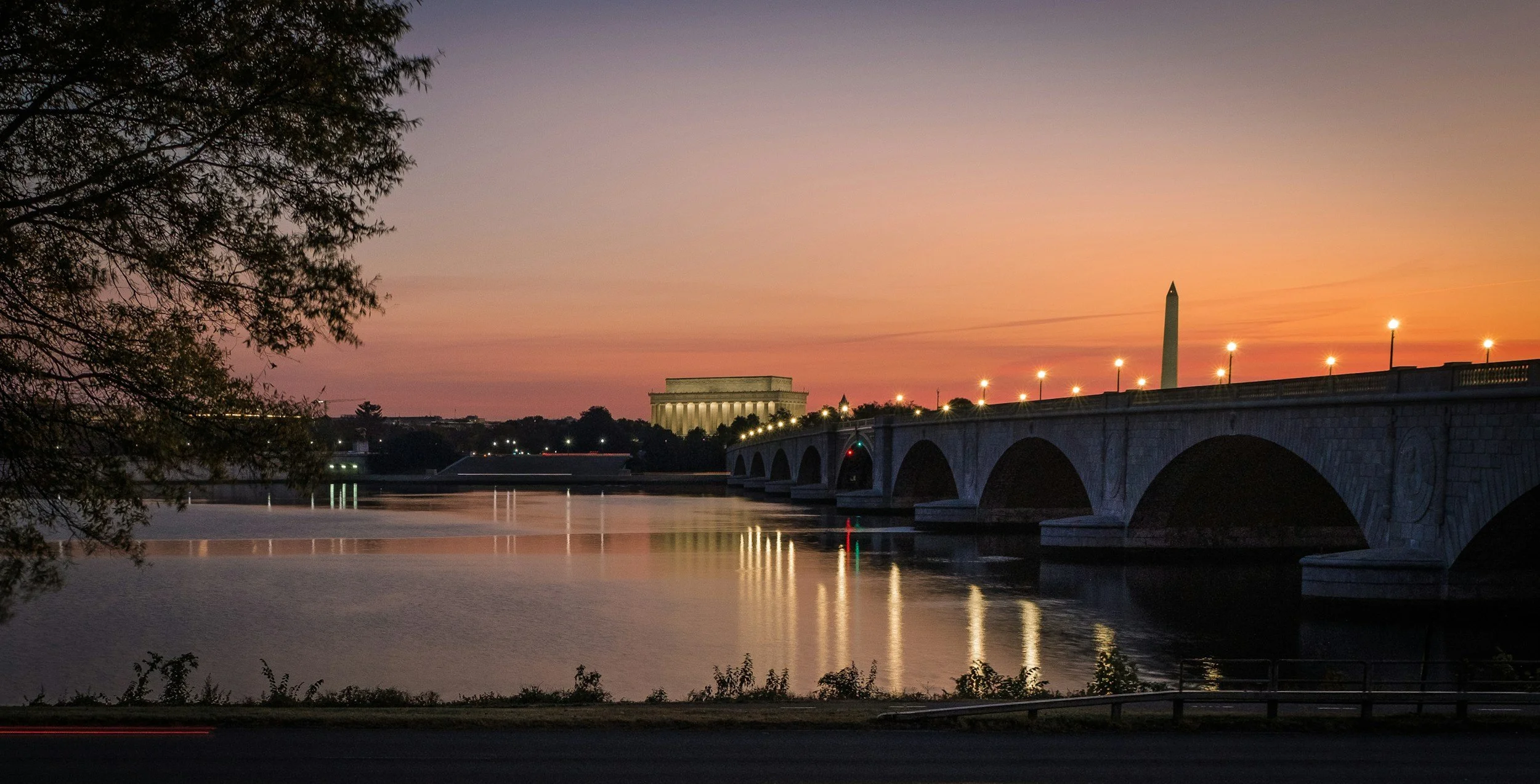A bridge over a river at sunset with the Lincoln Memorial and Washington Monument in the distance.