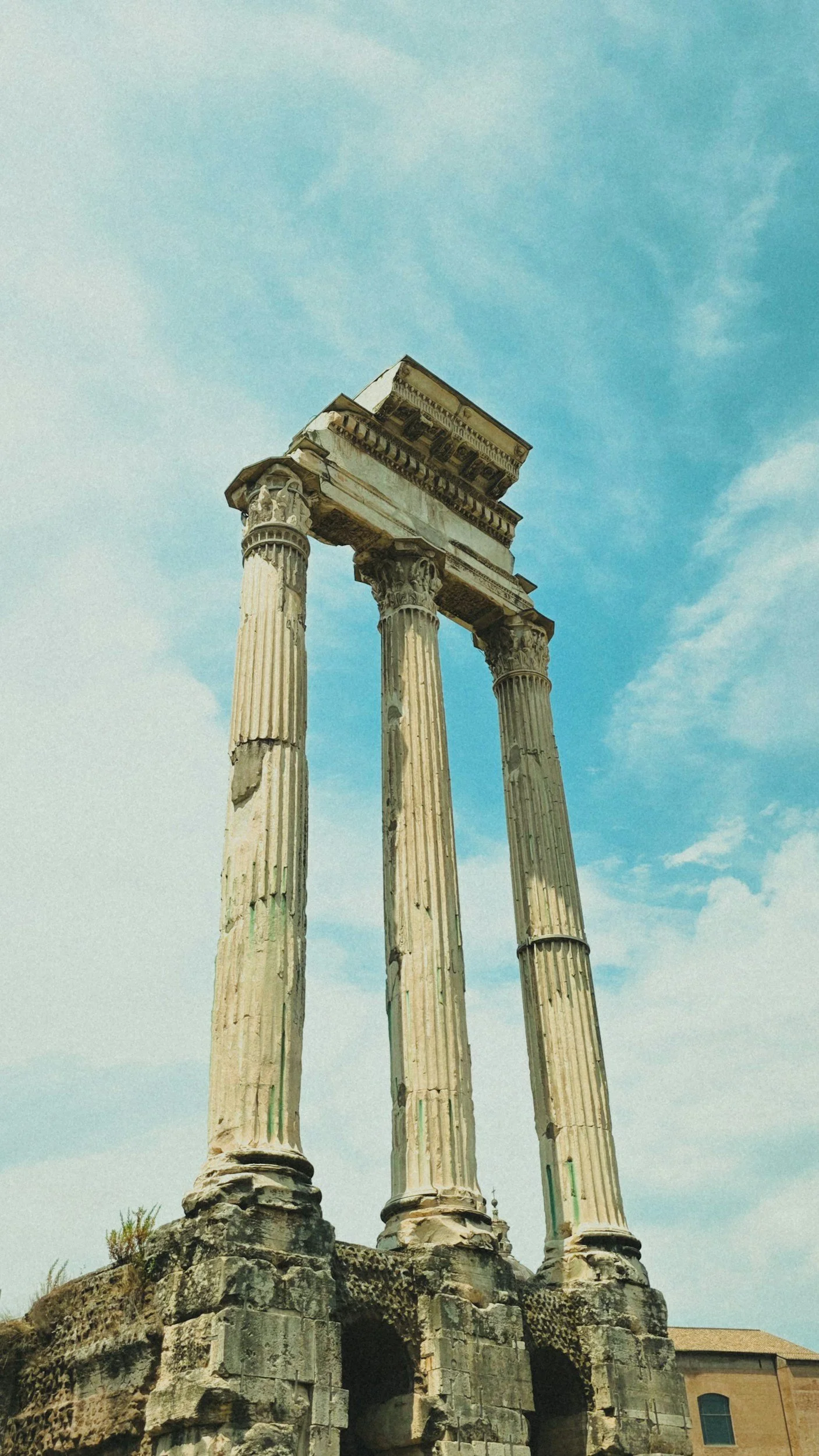 Ancient Greek ruins with three tall, weathered columns on a stone foundation, against a partly cloudy sky.