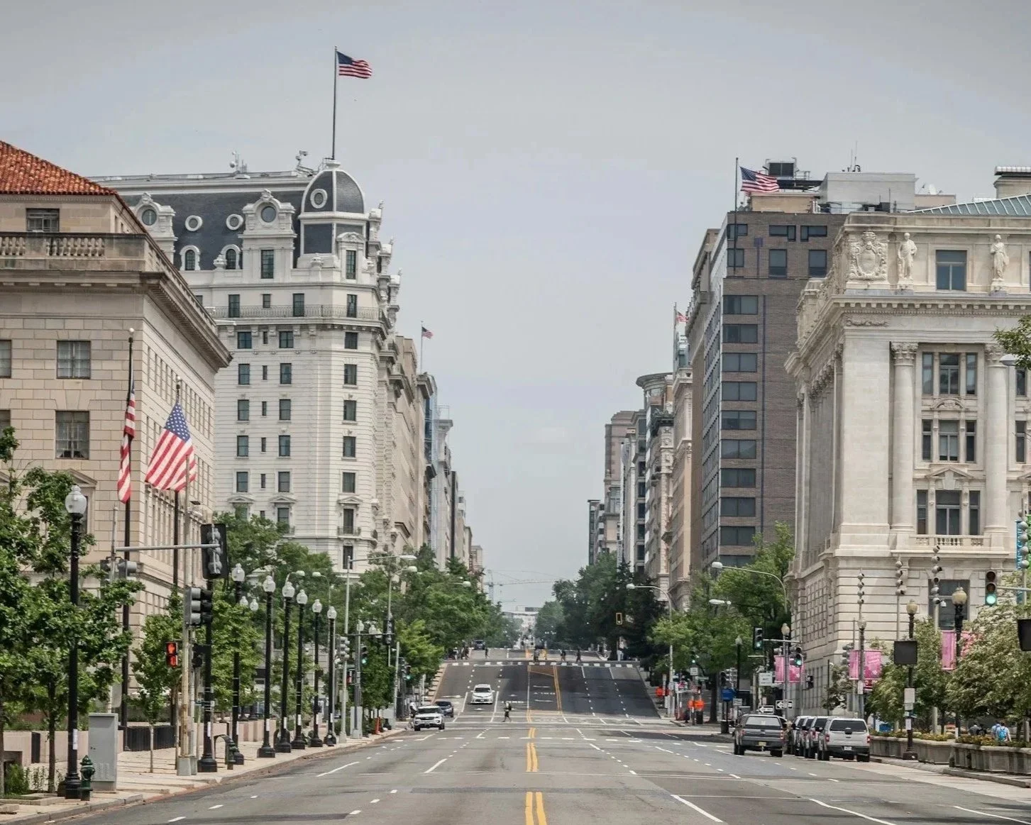 Empty city street with American flags, tall historic buildings, trees lining the sidewalks, and traffic lights at an intersection.