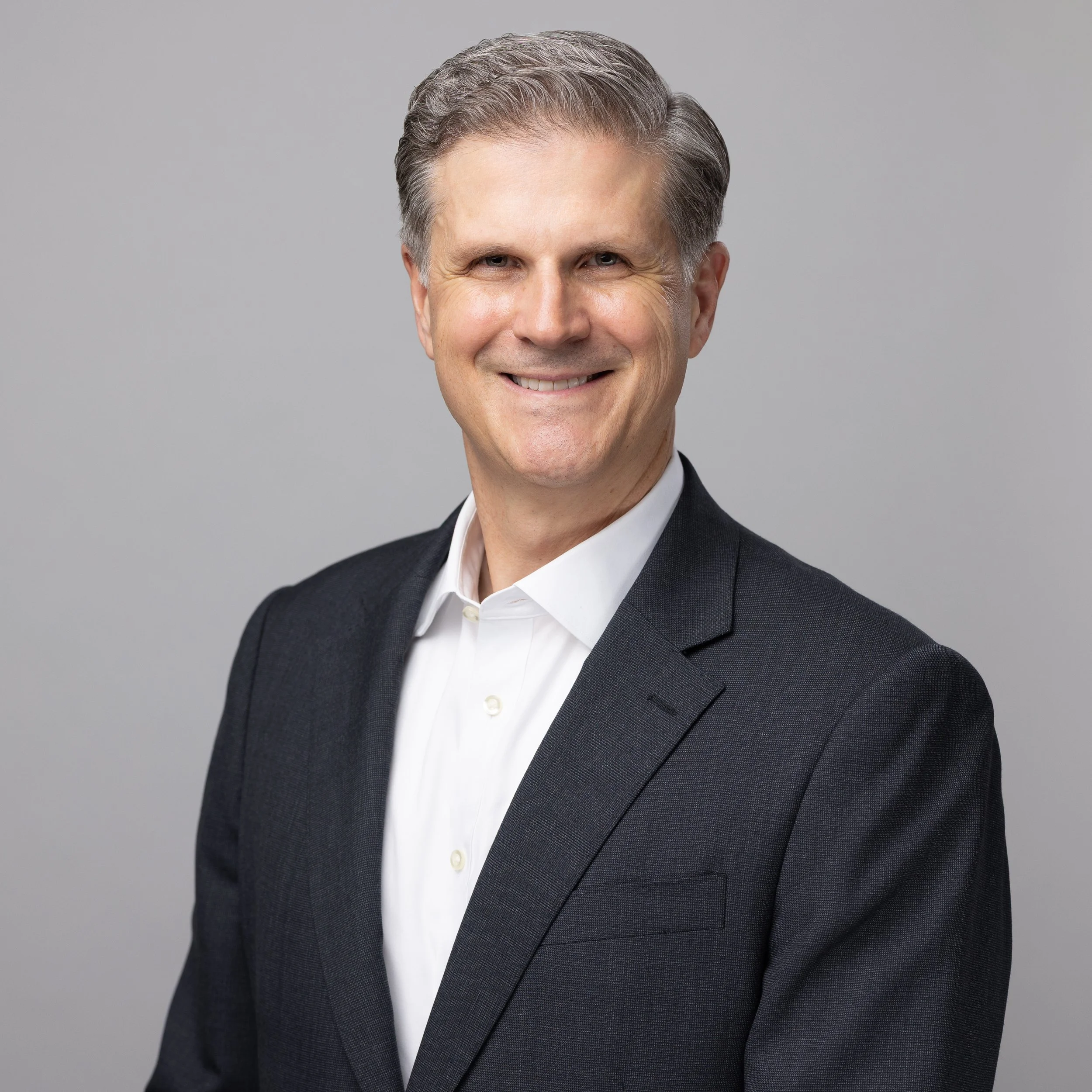 Close-up portrait of a middle-aged man with gray hair, smiling, wearing a dark suit and white shirt, against a plain gray background.