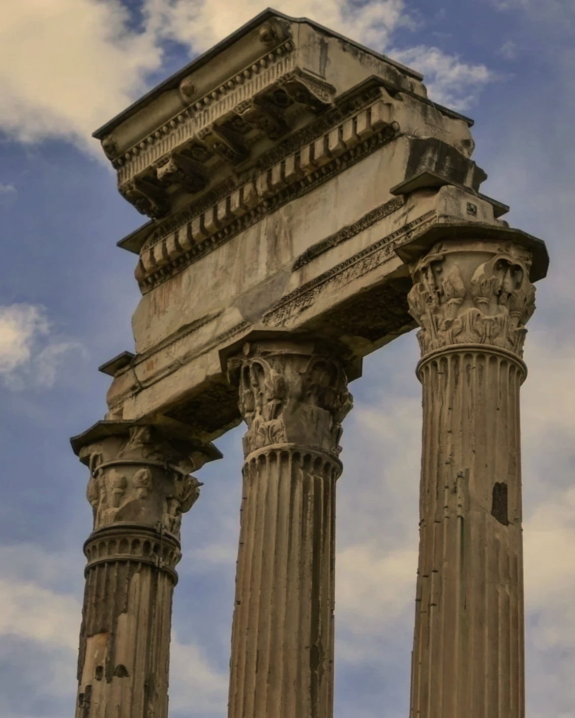 Close-up of ancient Greek ruin with three tall, fluted columns with carved Corinthian capitals and a partially preserved entablature against a partly cloudy sky.