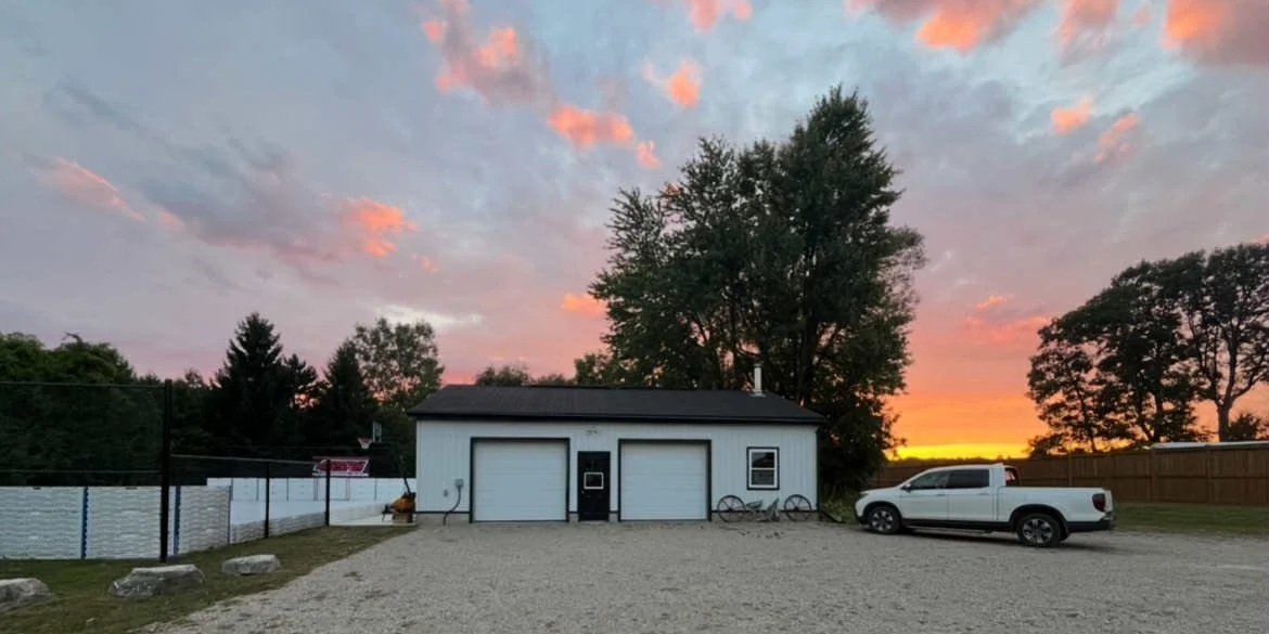 A small white building with two garage doors and a black door, set against a sunset sky with pink and orange clouds, with a white pickup truck and bicycles parked outside.