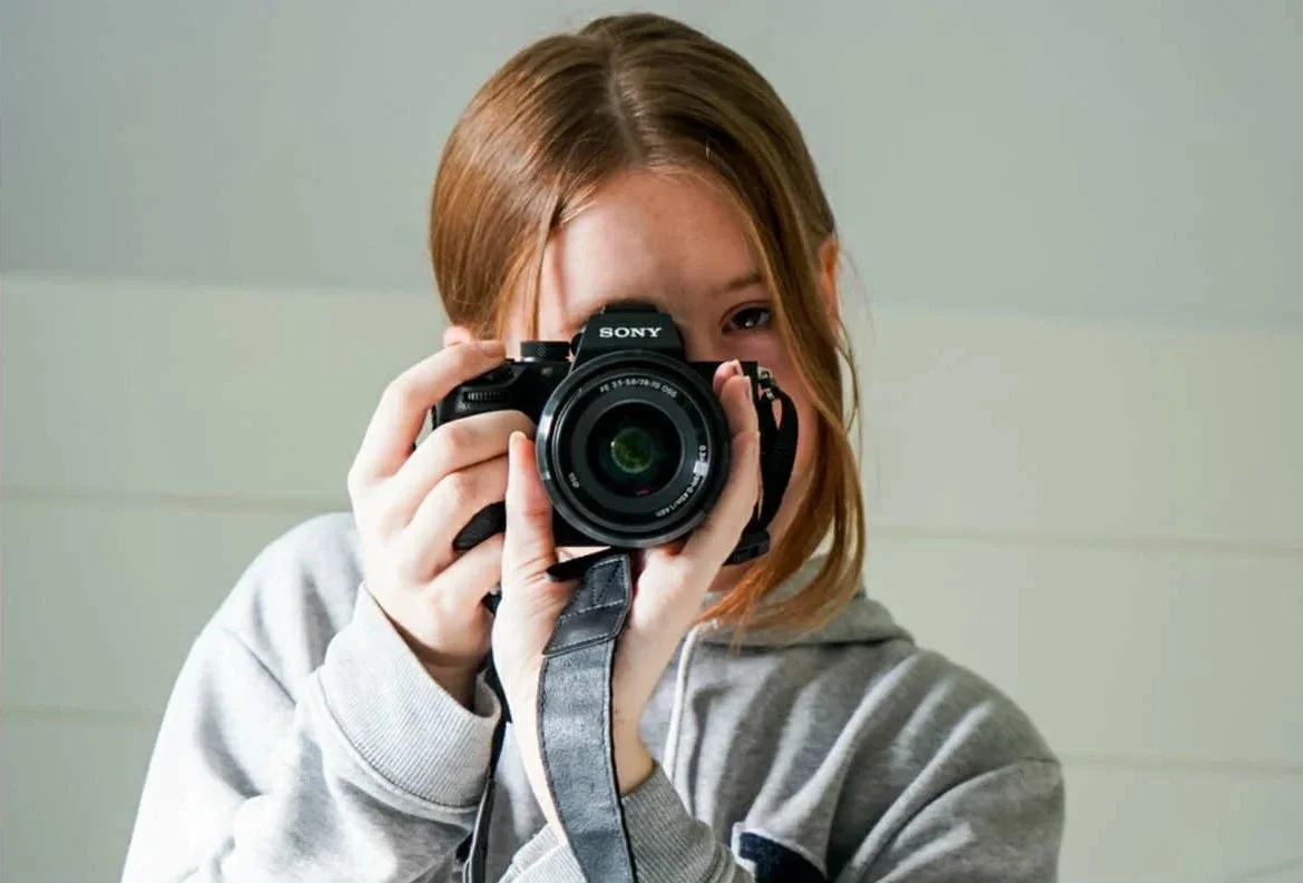 A young woman with reddish-brown hair taking a photo with a black Sony camera, wearing a gray hoodie, in front of a light-colored background.