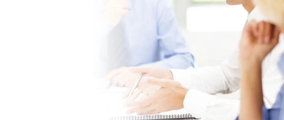 People sitting at a table in a meeting or discussion, one is taking notes with a pen.