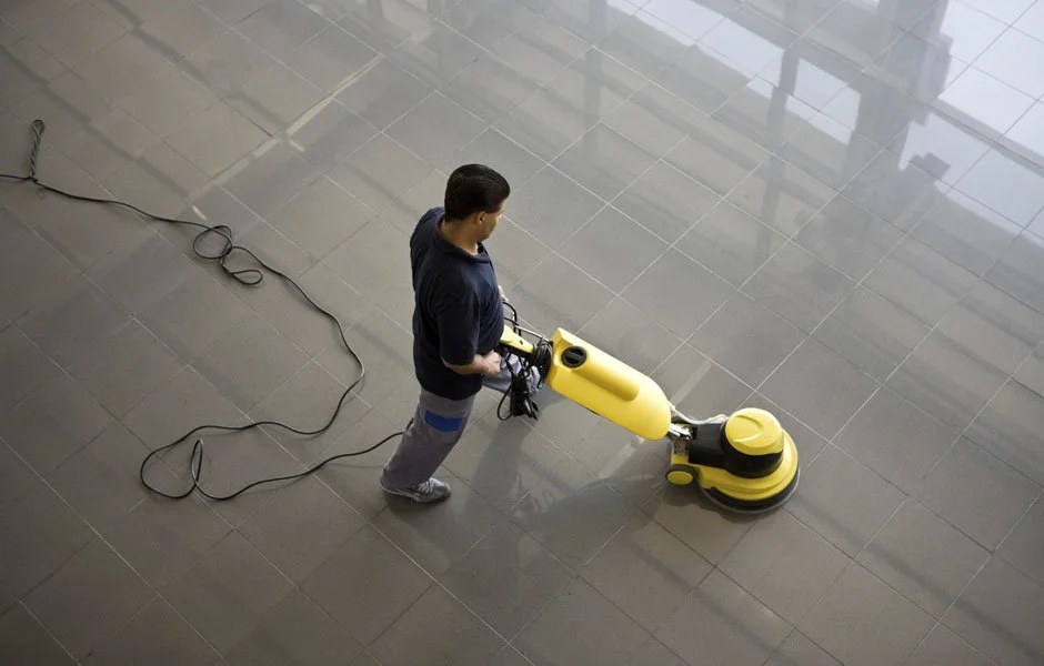 A man vacuuming a large, shiny tiled floor in a spacious, well-lit room.