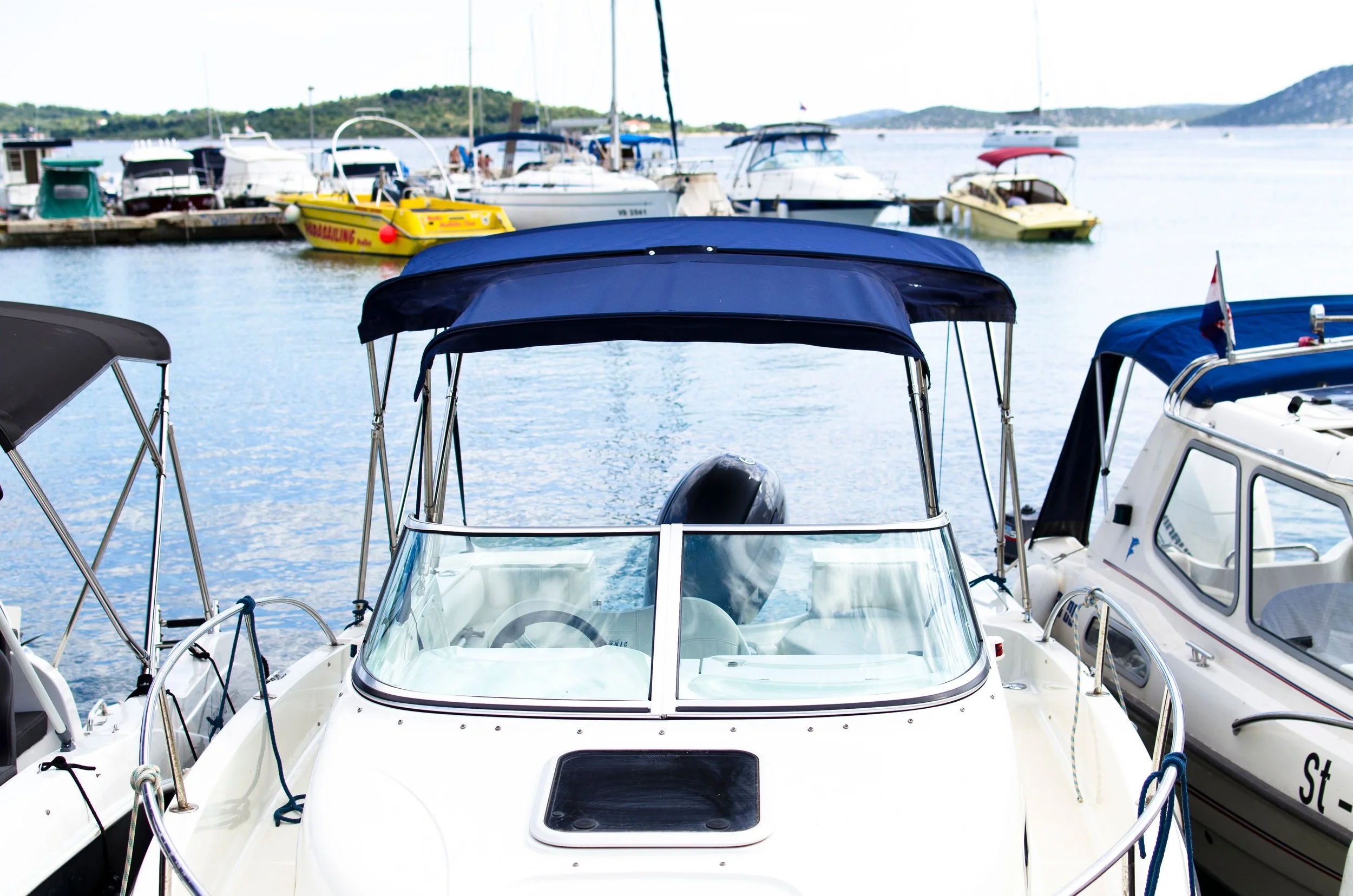 Multiple boats docked at a marina with water and hills in the background.
