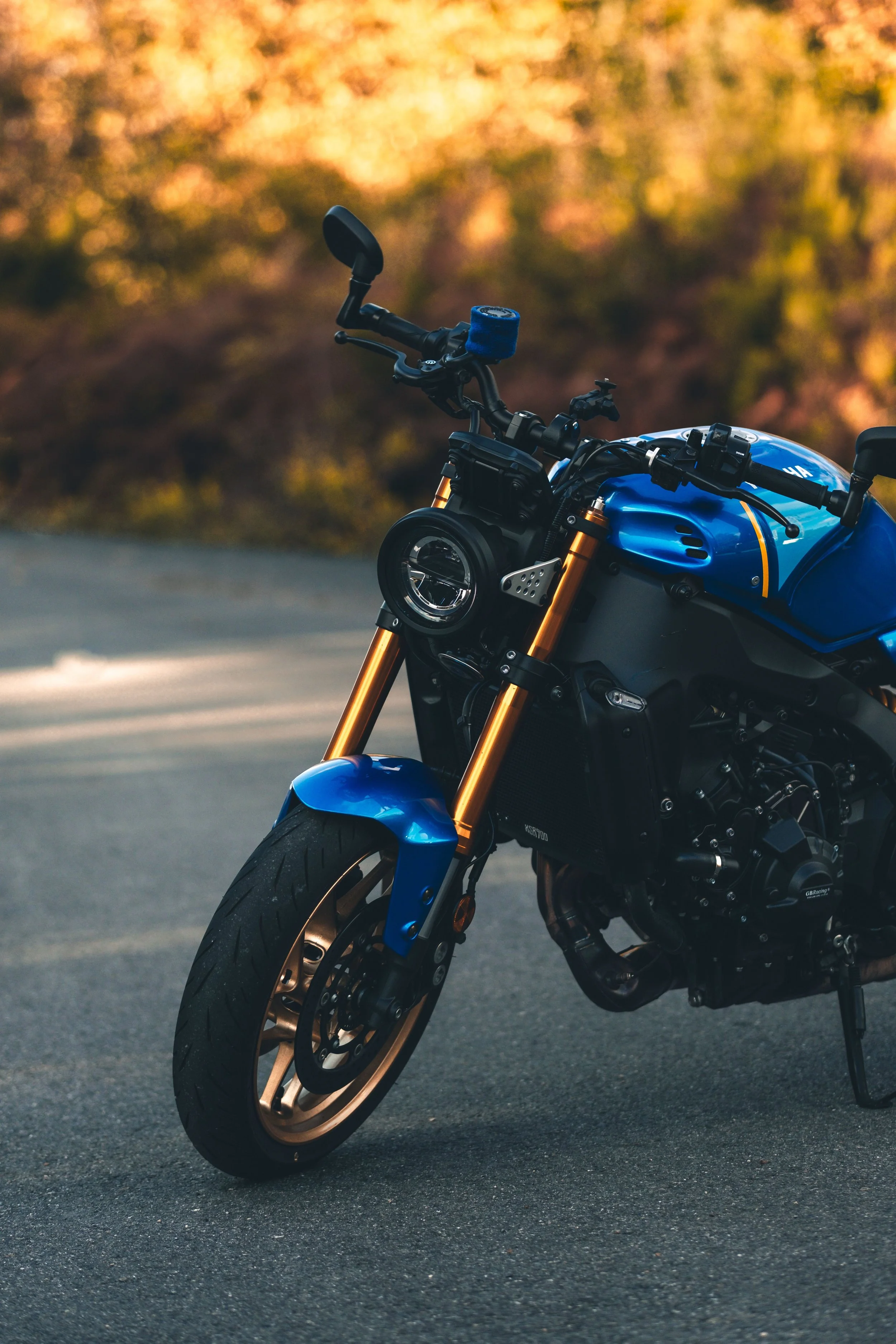 Close-up of a blue motorcycle with gold-colored suspension forks on an empty road, with a blurred background of trees in fall colors.