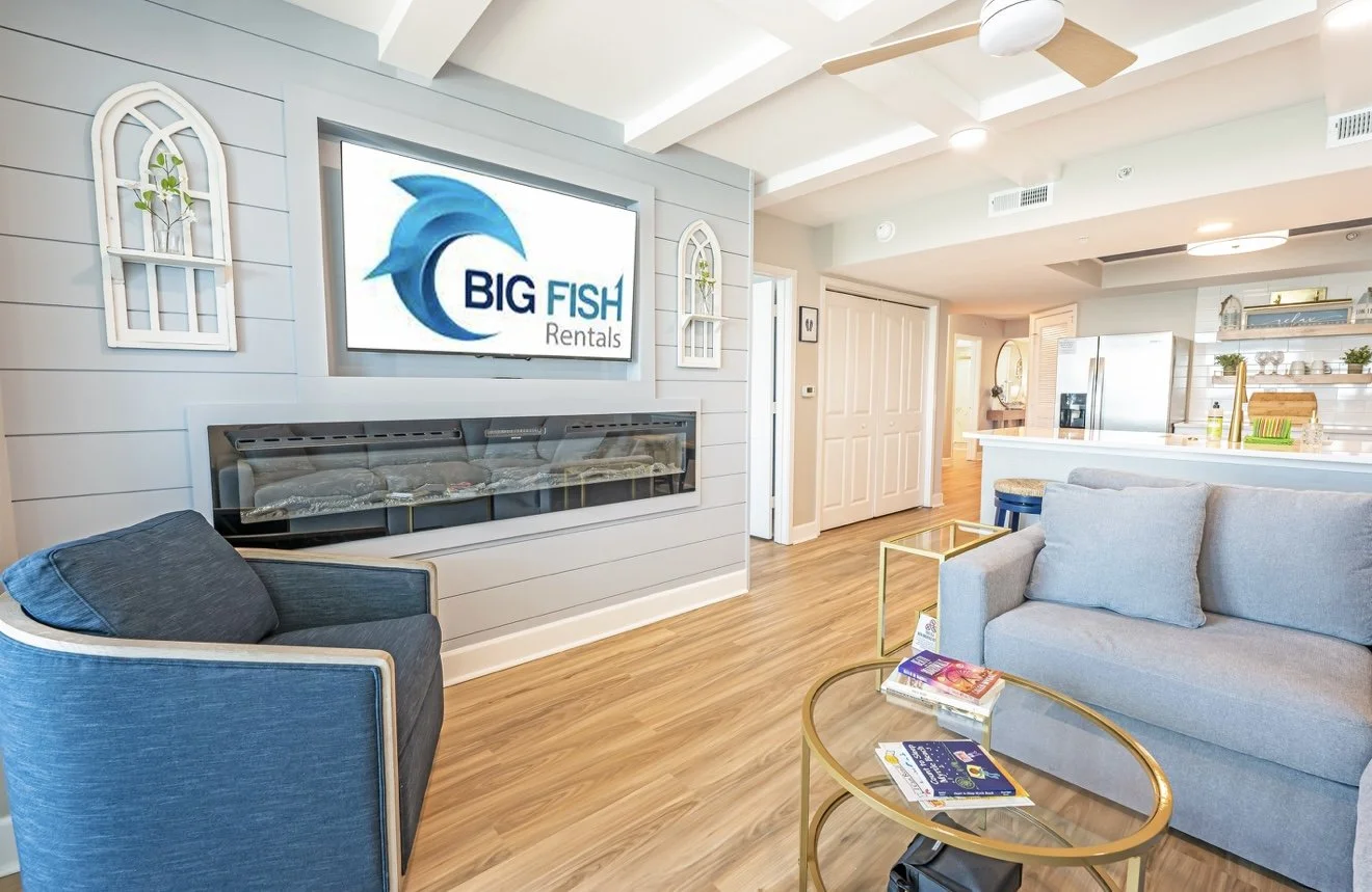 Sala de estar de uma casa moderna com cadeiras azuis e cinza, mesa de vidro com livros, televisão com logotipo de 'Big Fish Rentals', decoração minimalista com pequenas janelas decorativas, cozinha ao fundo com geladeira e prateleiras com utensílios e plantas.