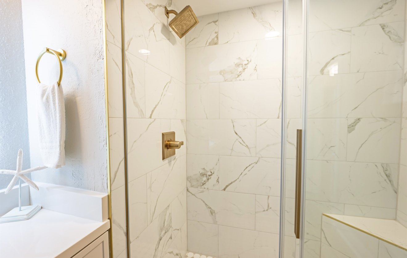 Banheiro com paredes de mármore branco, espaço de banho com chuveiro com cabeçada dourada e controle de água dourado, porta de vidro com puxador dourado, toalheiro dourado com toalha branca e decoração de estrela do mar na bancada