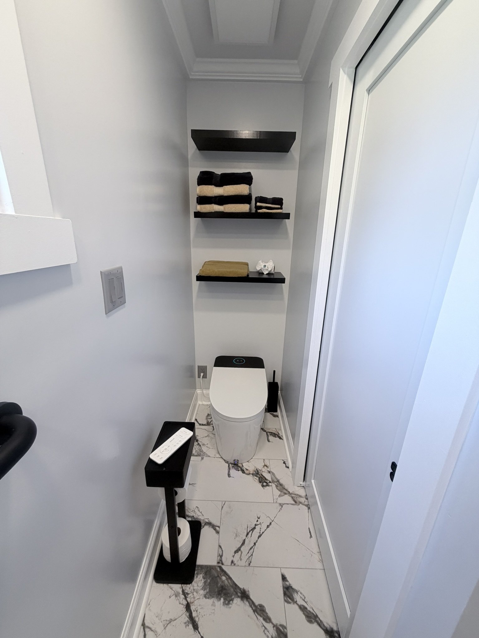 Banheiro moderno com piso de mármore branco com veios pretos, contendo um vaso sanitário, prateleiras negras com toalhas organizadas, uma mesa pequena com controle remoto e um papel higiênico, paredes brancas e uma porta de correr.