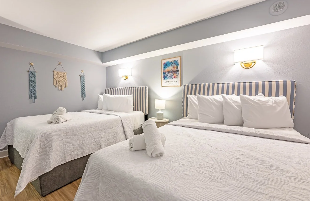 Quarto de hotel com duas camas de casal, cabeçalhos com listras azuis e brancas, paredes em tom claro, decorada com quadro e tecelagens na parede, luzes de parede, luminária de cabeceira acesa, toals de banho dobrados no centro das camas.