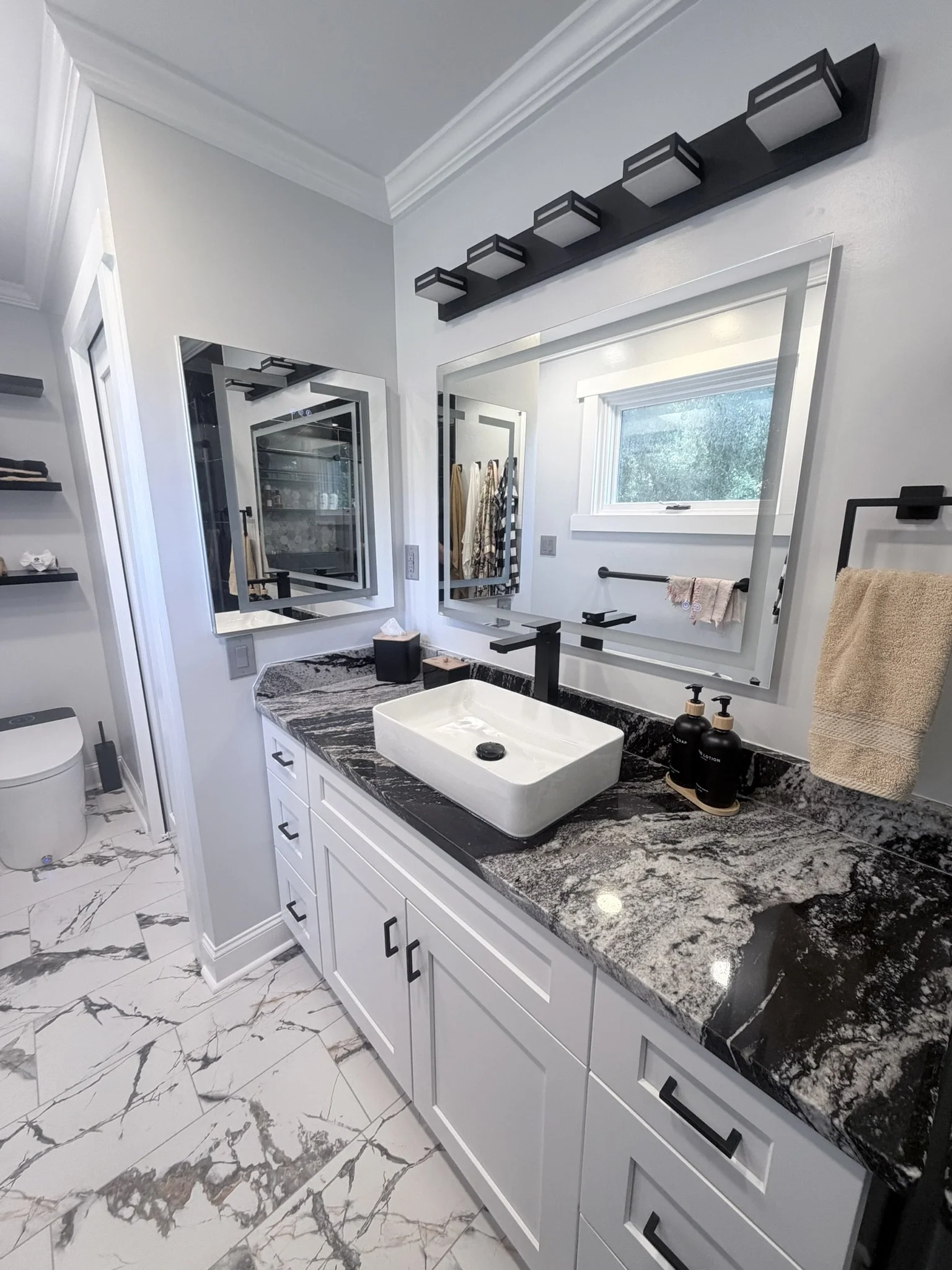 Banheiro moderno com pia de bancada de granito preto e branco, espelhos grandes acima da pia, armários brancos com tampo de granito, piso de mármore branco com veios cinza, janela com vista para árvores, toalhas penduradas em barra, produtos de higiene no balcão