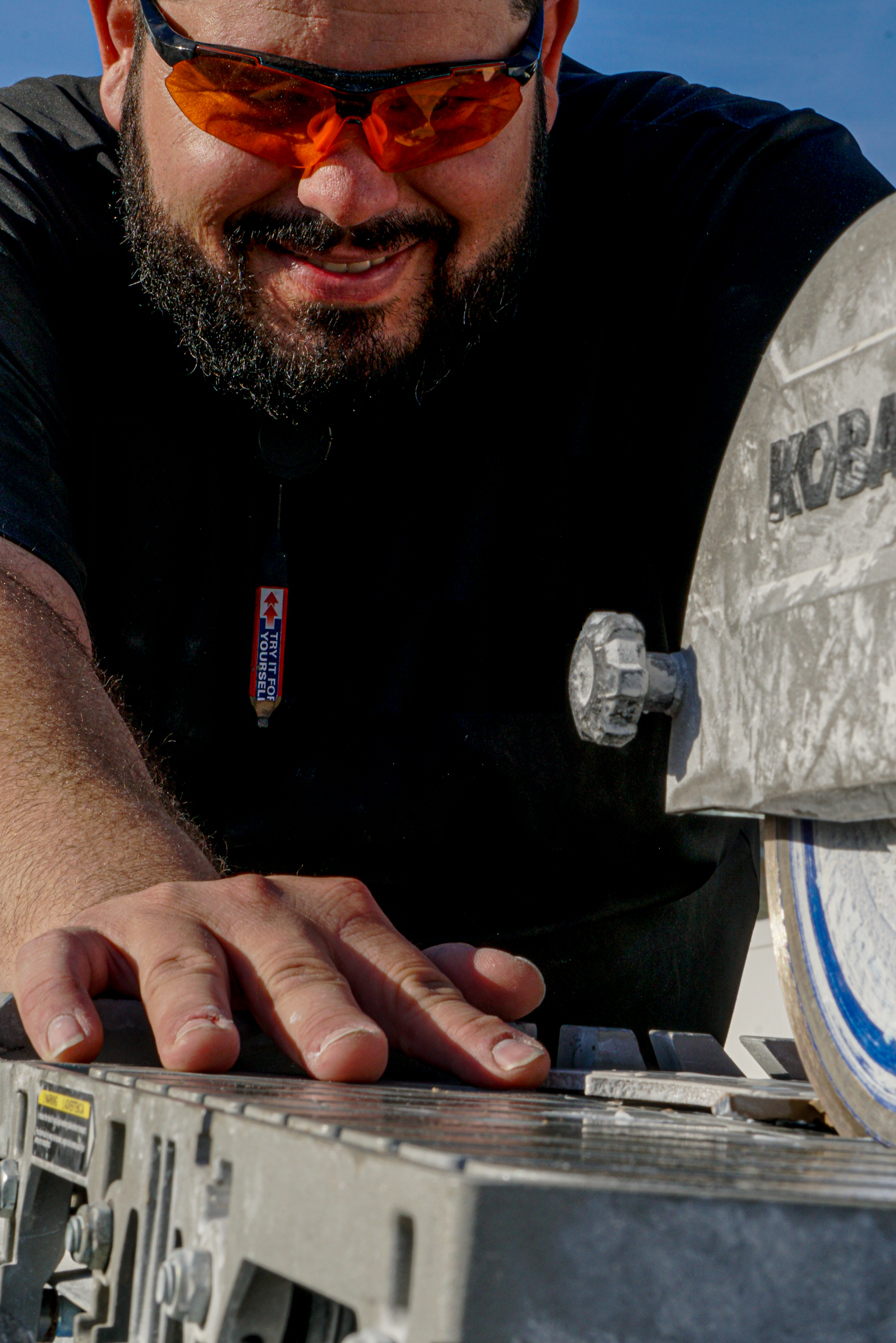 Homem usando óculos de proteção trabalhando com equipamento de metal, segurando uma peça com as mãos em um ambiente de oficina ou construção.