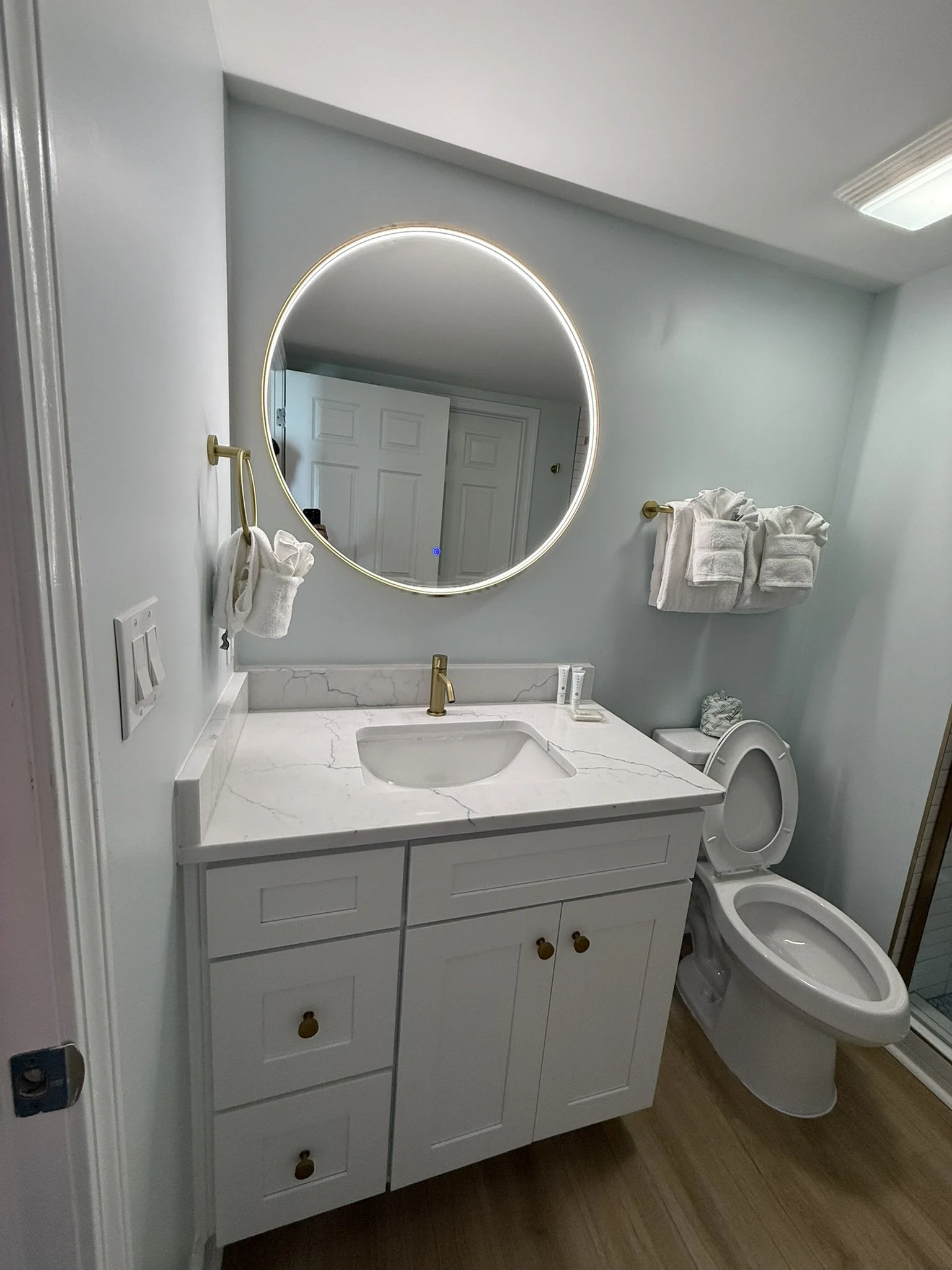 Banheiro com espelho arredondado com iluminação LED, pia de mármore branco, armário branco com portas e gavetas, toalhas brancas, vaso sanitário e piso de madeira.