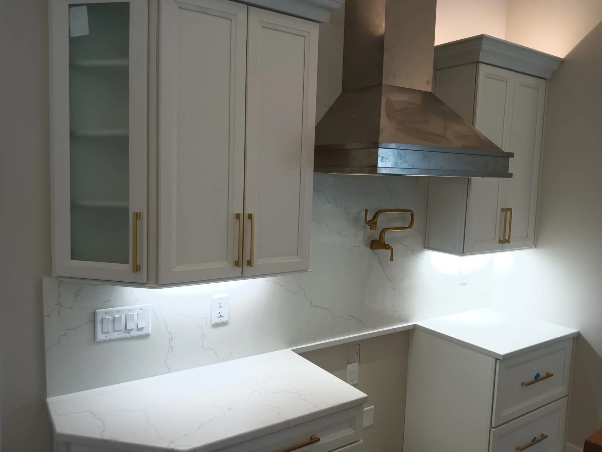 Cozinha moderna com armários brancos, bancada de mármore branco com veios cinza, e uma coifa de aço inox. Há tomadas na parede e uma torneira dourada em uma pia de cozinha.