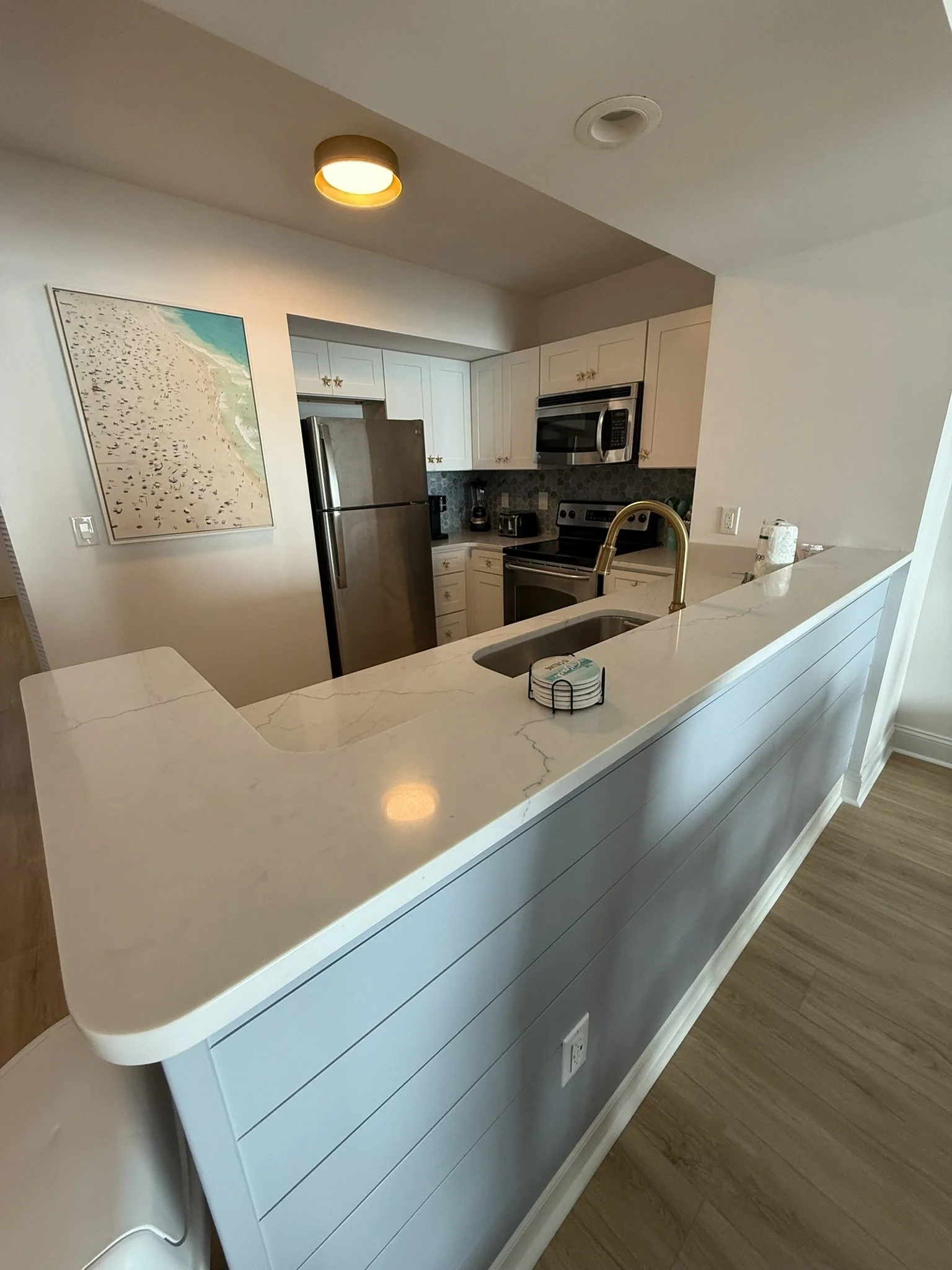 Vista de uma cozinha moderna com armários brancos, pia com torneira dourada, eletrodomésticos de inox como geladeira, forno micro-ondas, fogão e utensílios na bancada. Uma parede decorada com azulejos hexagonais e uma pintura de praia na parede ao lado.