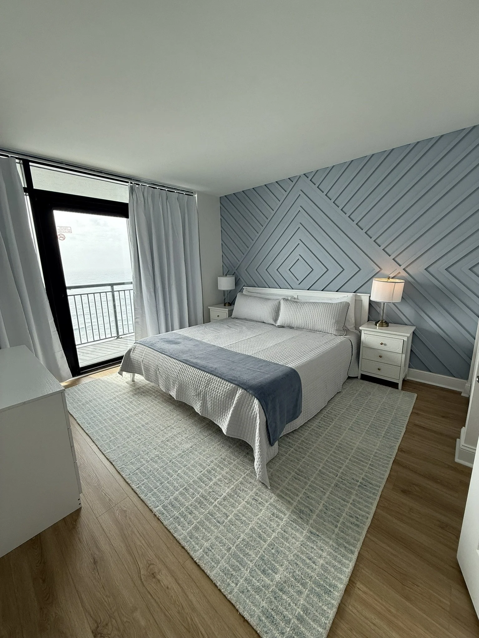 Quarto de hotel com cama de casal, almofadas, dois criados-mudos com abajures, tapete claro, cortinas brancas, cerca de uma janela com vista para o mar