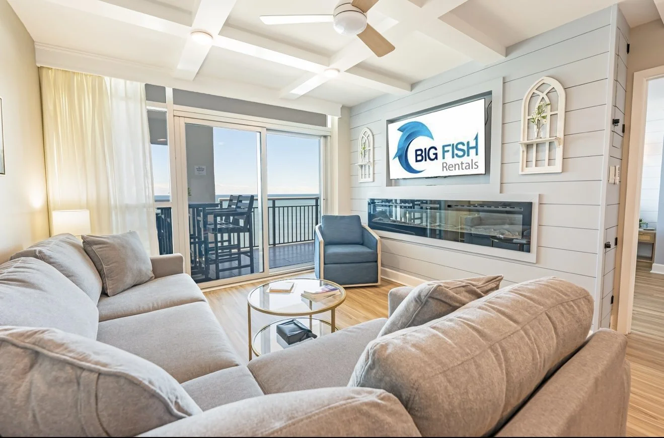 Sala de estar decorada com sofá bege, poltrona azul, mesa de centro de vidro, televisão de tela plana com logo Big Fish Rentals, parede branca de Painel de madeira, cortinas brancas, porta de vidro com vista para varanda com vista para o mar.