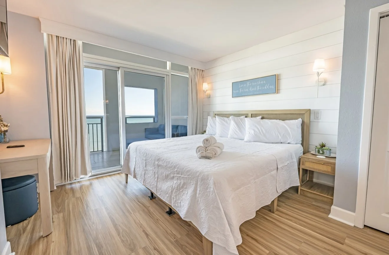 Quarto de hotel com cama de casal, almofadas brancas, toalhas enroladas sobre a cama, varanda com cadeira azul e vista para o oceano, decoração clara e aconchegante.