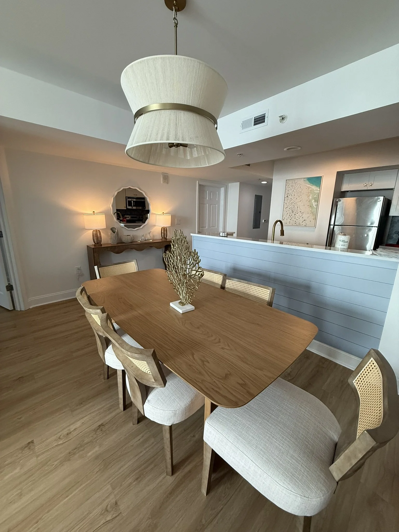 Sala de jantar com mesa de madeira retangular, cadeiras com encosto de cana e almofada clara, luminária de teto grande, espelho decorativo na parede ao fundo, e cozinha ao lado com geladeira de inox e pintura de paisagem na parede.