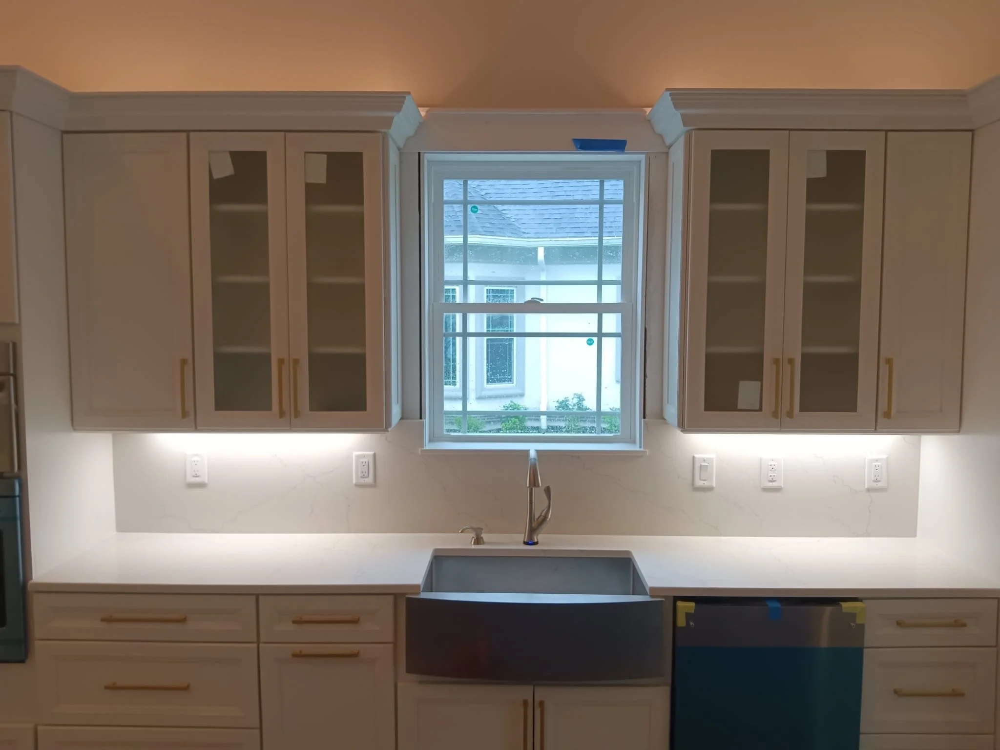 Cozinha moderna com armários brancos, bancadas de mármore branco, pia de inox, janela de vidro grande com vista para o exterior e luzes embutidas no teto.