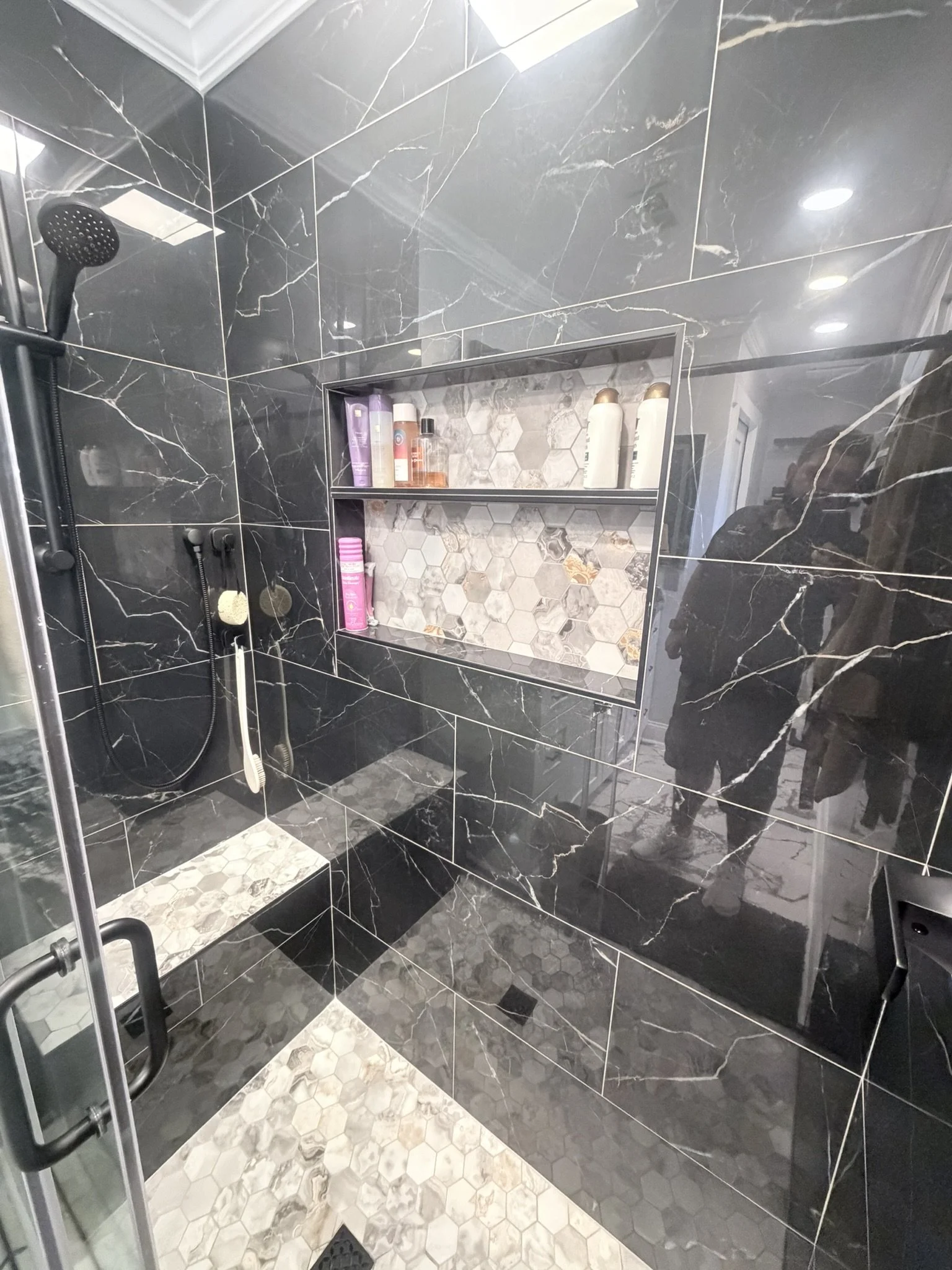 Banheiro com paredes e teto revestidos de azulejos pretos com veios brancos, nicho embutido com revestimento em mosaico de hexágonos em tons de cinza e detalhes dourados, prateleira de vidro para produtos de higiene, box de vidro com porta de metal preto, chão com azulejos hexagonais em tons de cinza e branco, banheira, chuveiro e acessórios de banho.