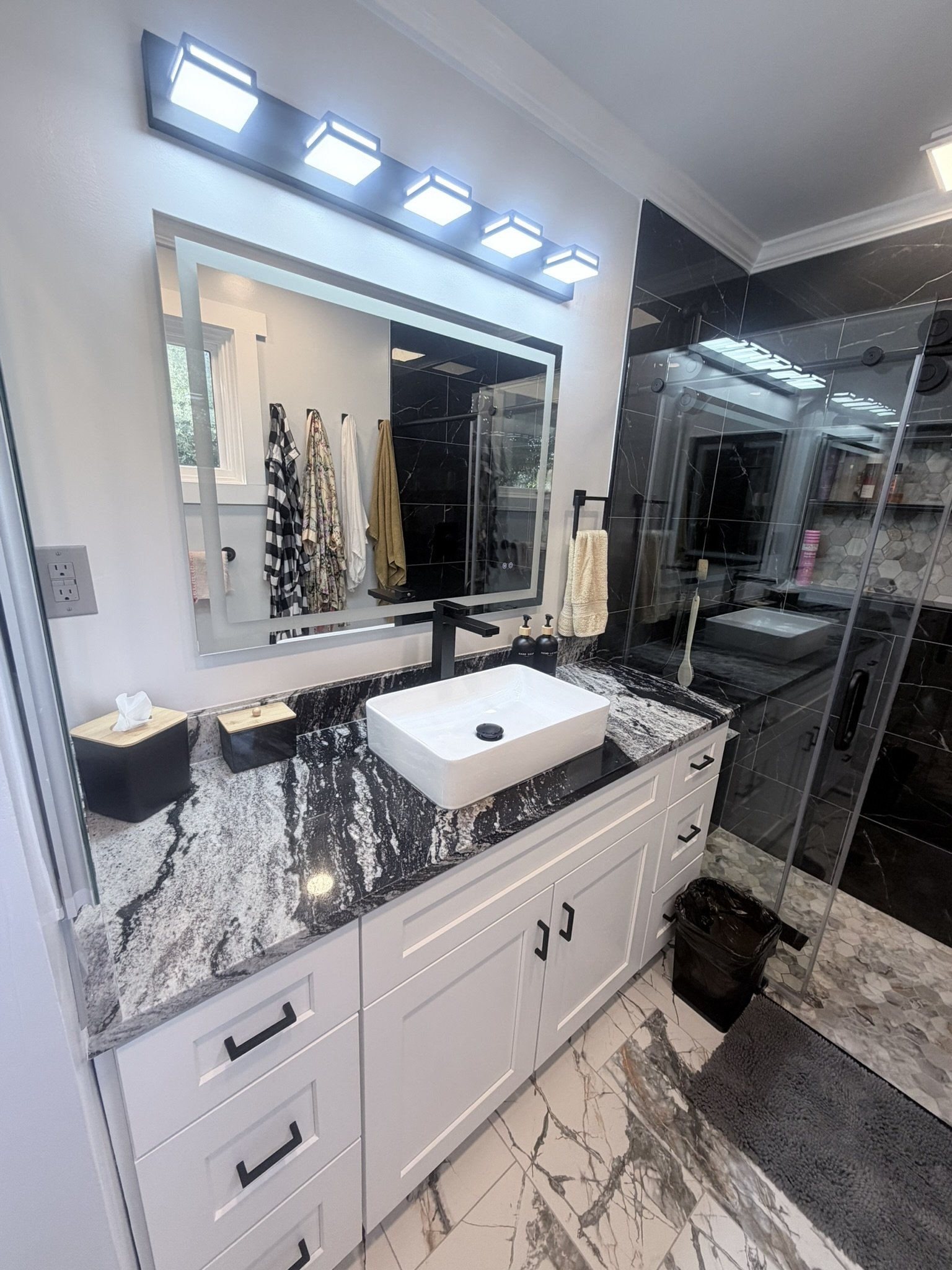 Banheiro moderno com pia de bancada de mármore preto e branco, espelho grande, iluminação de LED e detalhes em dourado. Toalhas penduradas, produtos de higiene na pia, portas de vidro de box, piso de mármore claro com veios escuros, tapete cinza escuro e lixo preto ao lado do vaso.