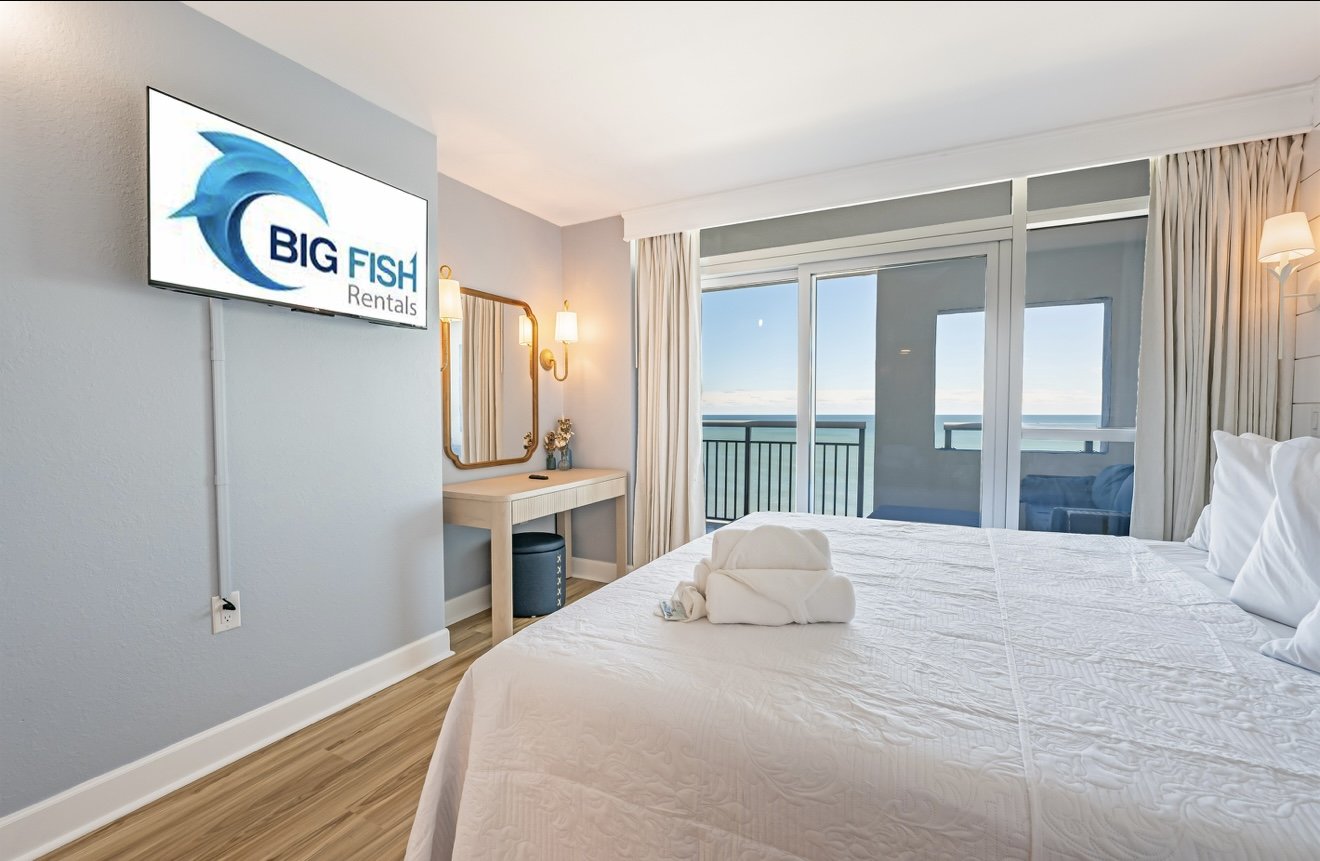 Quarto de hotel com vista para o mar, cama arrumada com toalhas dobradas, televisão na parede com o logo "Big Fish Rentals", cortinas claras, espelho e luminárias de parede.