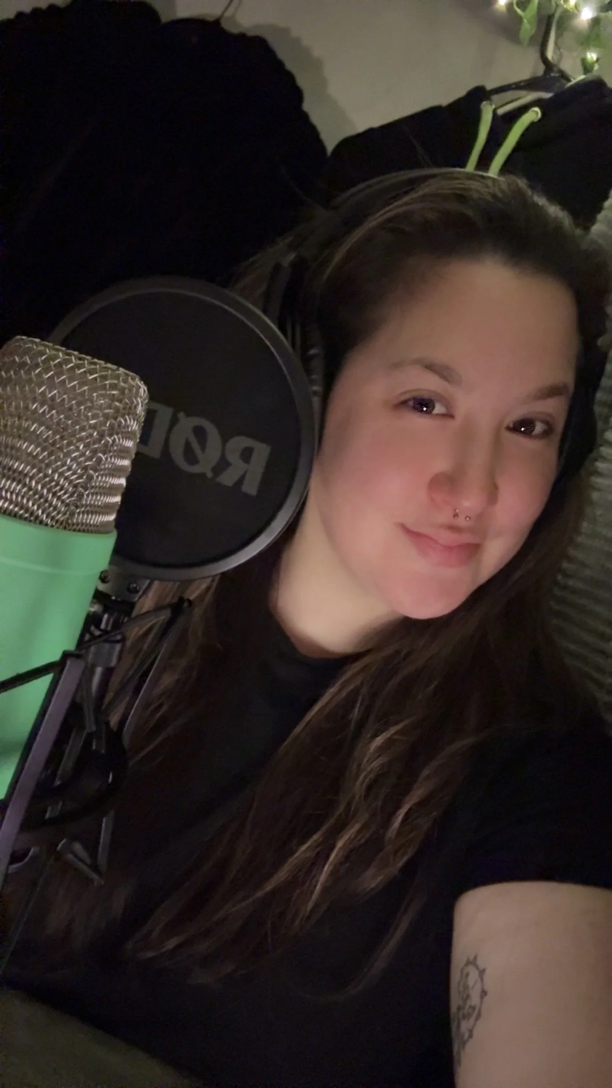 A young woman with long brown hair sitting behind her microphone, labeled "RODE". She has a nose ring and a tattoo on her left arm.
