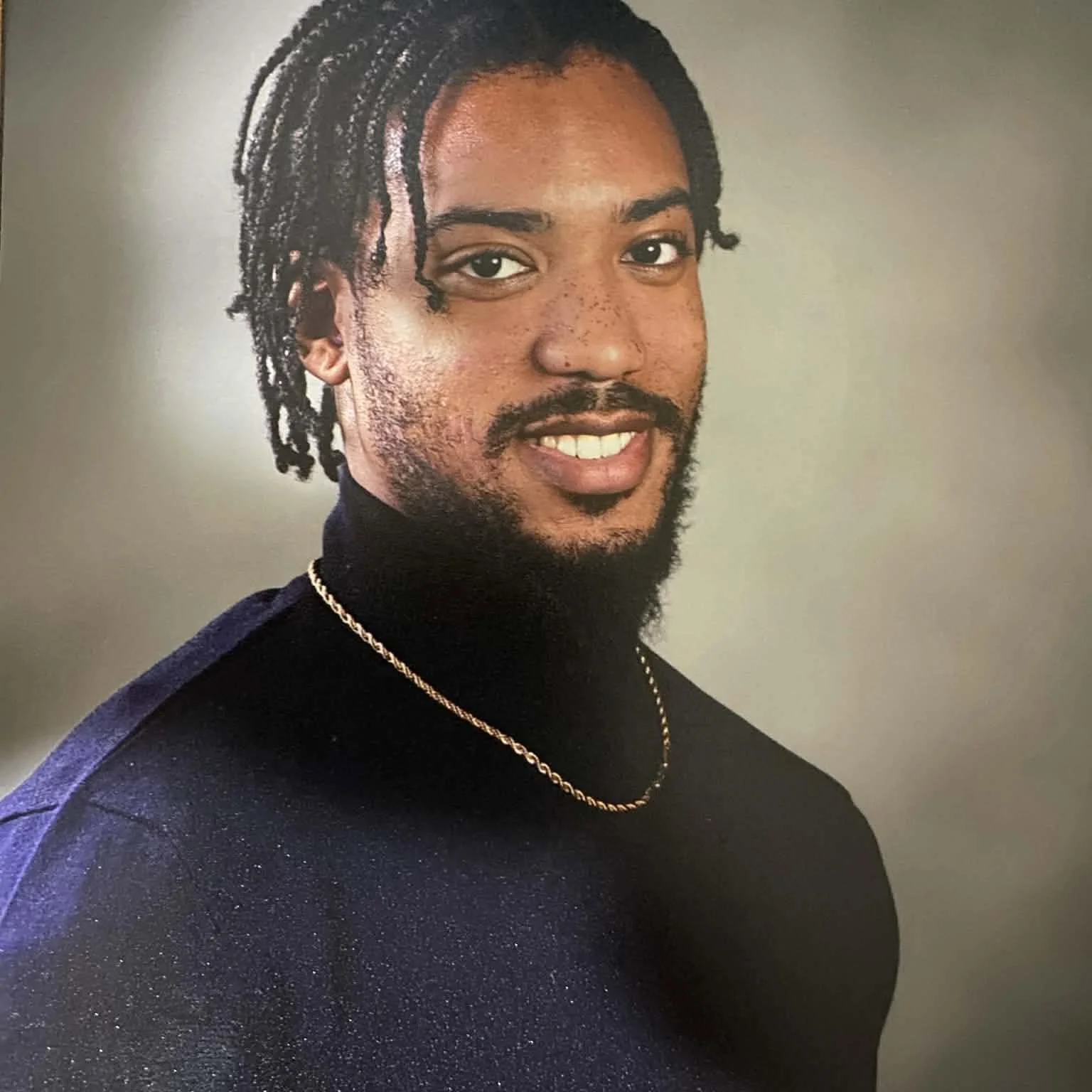A portrait of a man with dreadlocks, a mustache, and a goatee, wearing a black turtleneck and a gold chain necklace, smiling and facing the camera against a plain background.