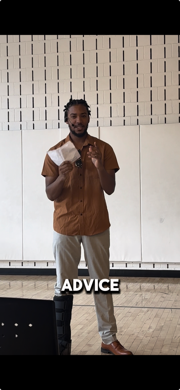 A man with dreadlocks wearing a brown shirt and beige pants giving advice during a presentation. He is holding a face mask in one hand and gesturing with the other. The setting appears to be indoors with a brick and plain wall backdrop. The word "ADVICE" is overlaid on the image.