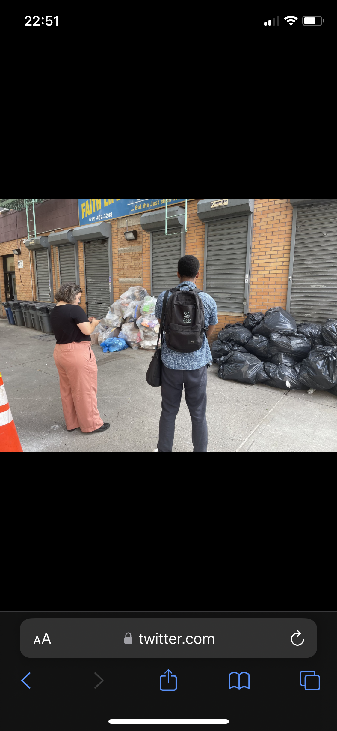Two people standing on a sidewalk near a pile of black garbage bags and other trash bags filled with waste, in front of a row of closed metal shutters on a brick building.