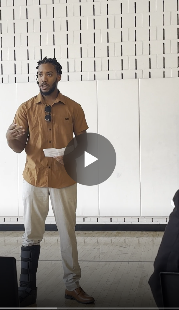 A man with dreadlocks, wearing a brown shirt, light-colored pants, and a black medical boot on his right foot, is standing and speaking with a paper in hand in front of a white brick wall.