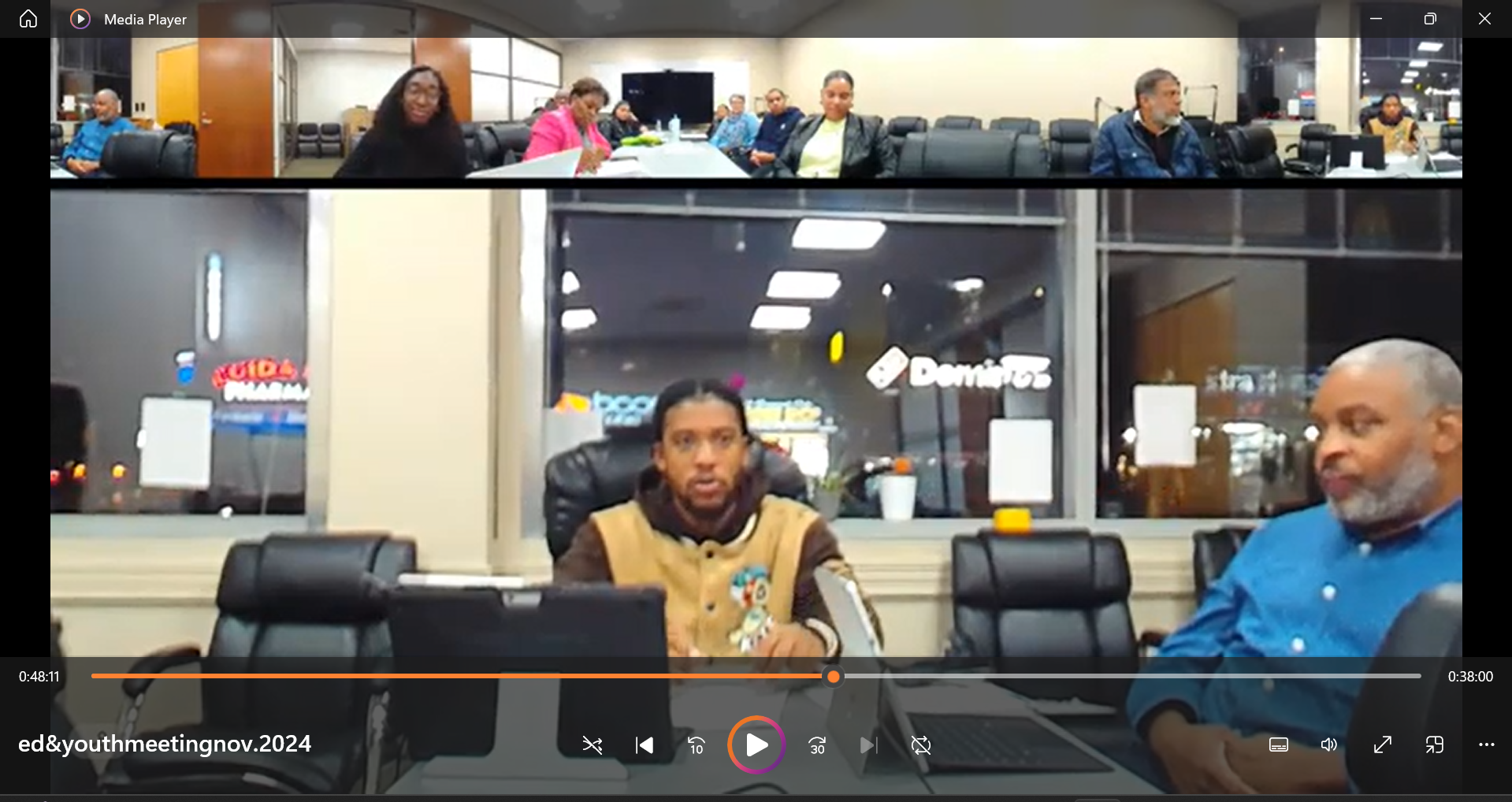 A group of diverse people sitting around conference tables in a meeting room, with some engaged in discussion and others listening, seen through a split-screen video call showing an indoor setting outside a store at night.