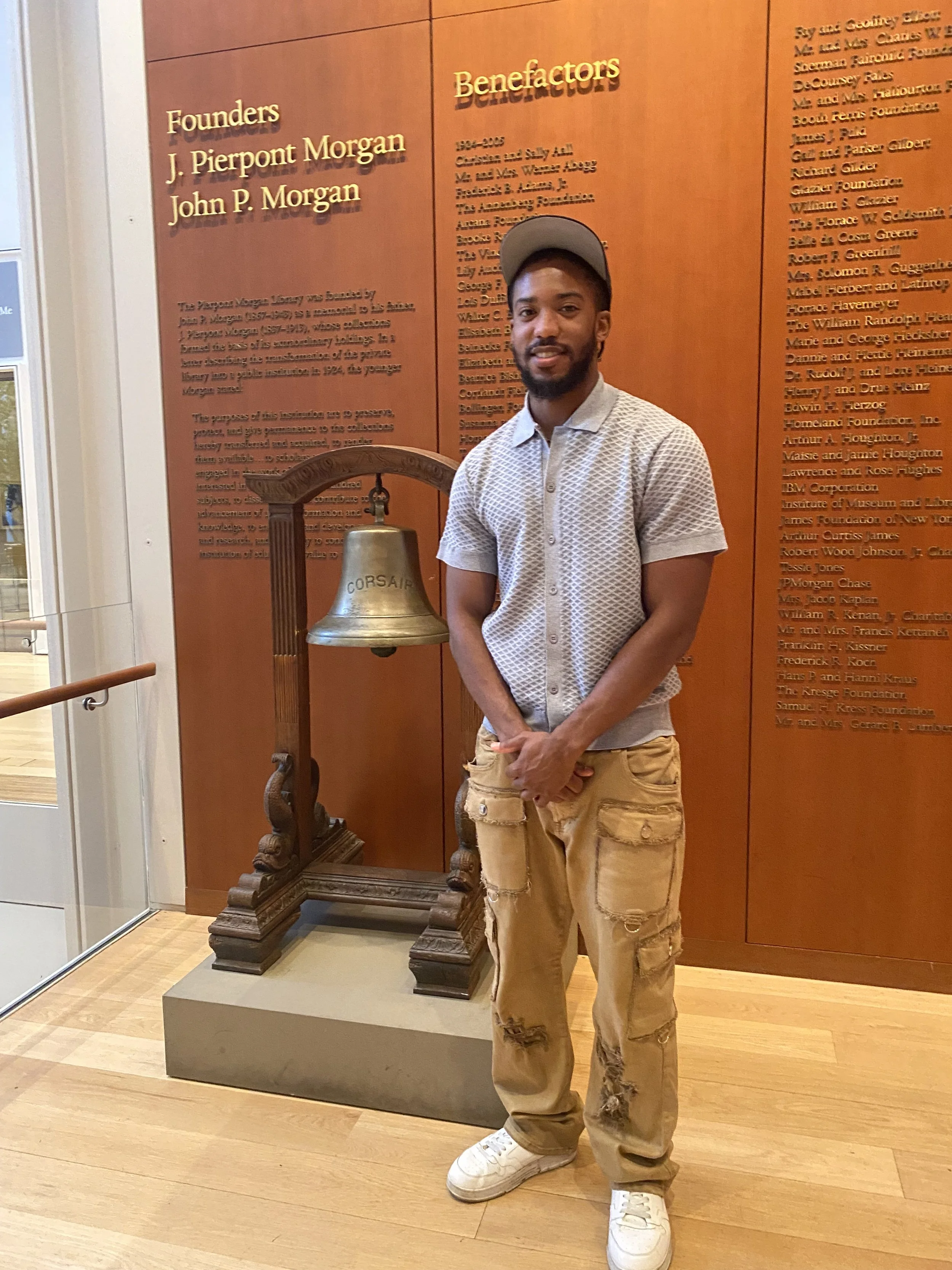 A young man with a beard and wearing a gray baseball cap, a light patterned polo shirt, and tan distressed cargo pants stands in front of a historical display featuring a large bronze bell on a wooden stand and a wall with gold-lettered informational text. The man is casually smiling at the camera.