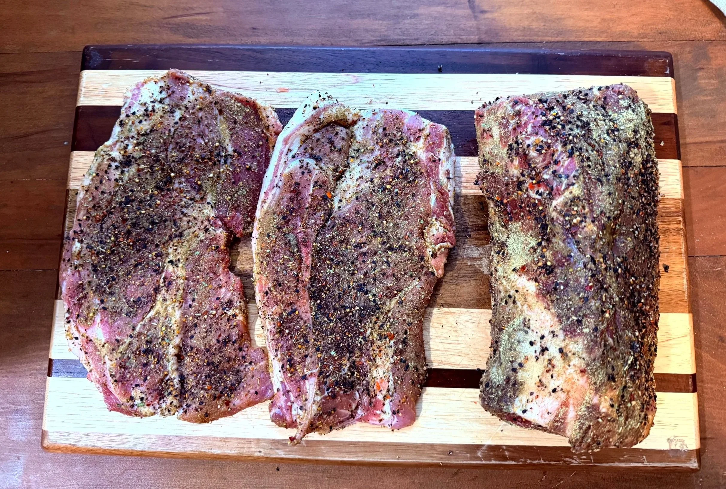 Three pieces of seasoned raw meat on a wooden cutting board.