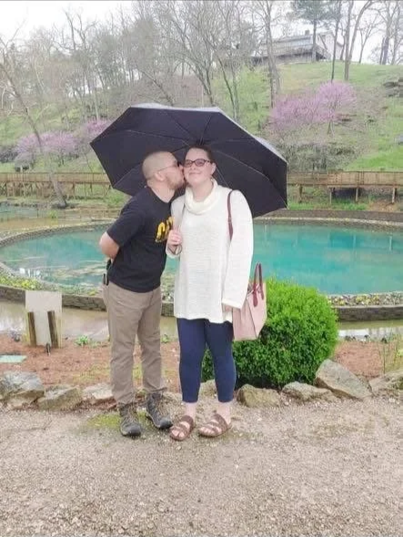 A man is kissing a woman on the cheek while standing close together under a black umbrella by a pond with blossoming trees in the background.