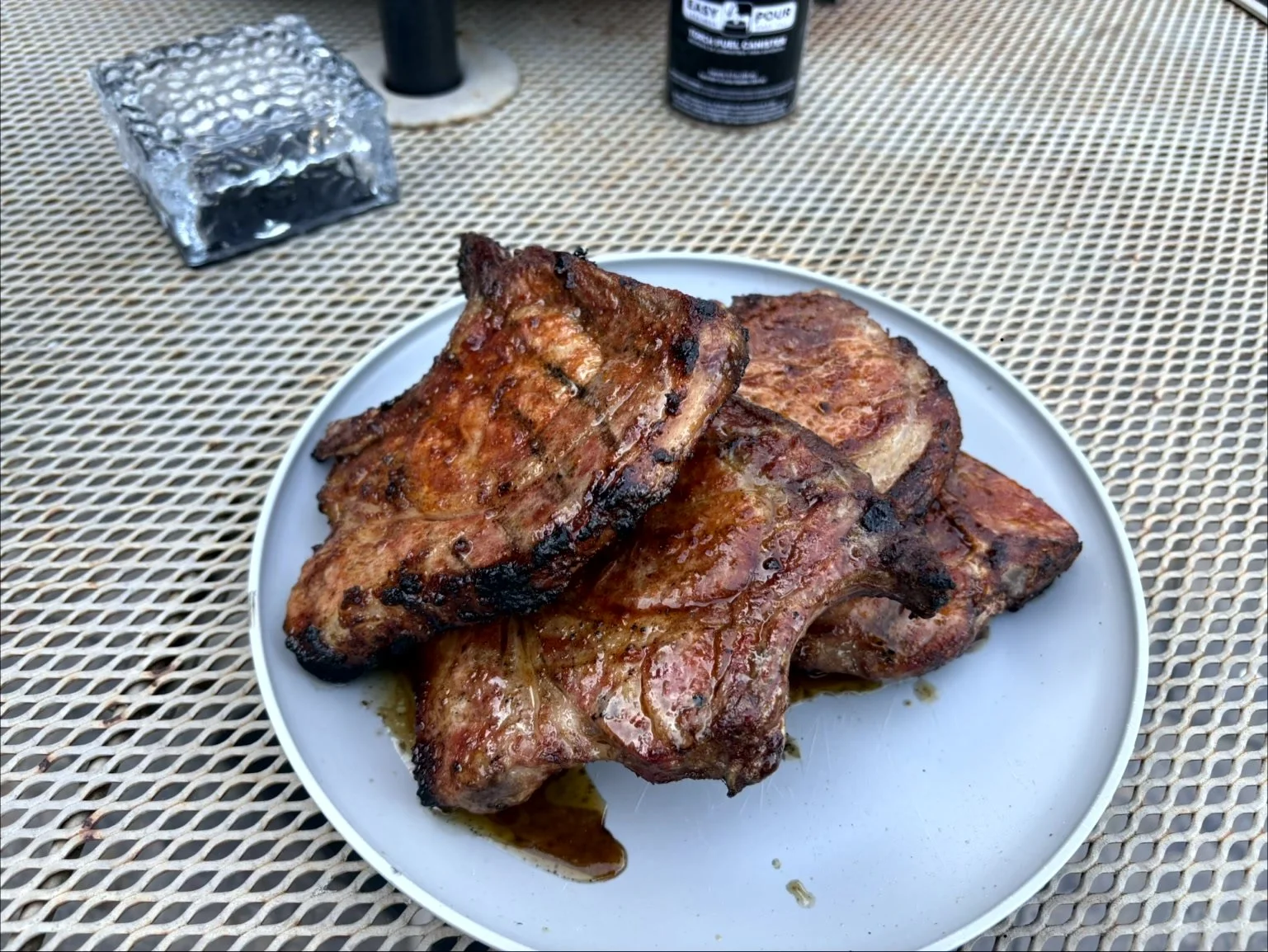 Close-up of grilled pork steaks on a white plate, placed on a metal mesh table with condiments and utensils in the background.