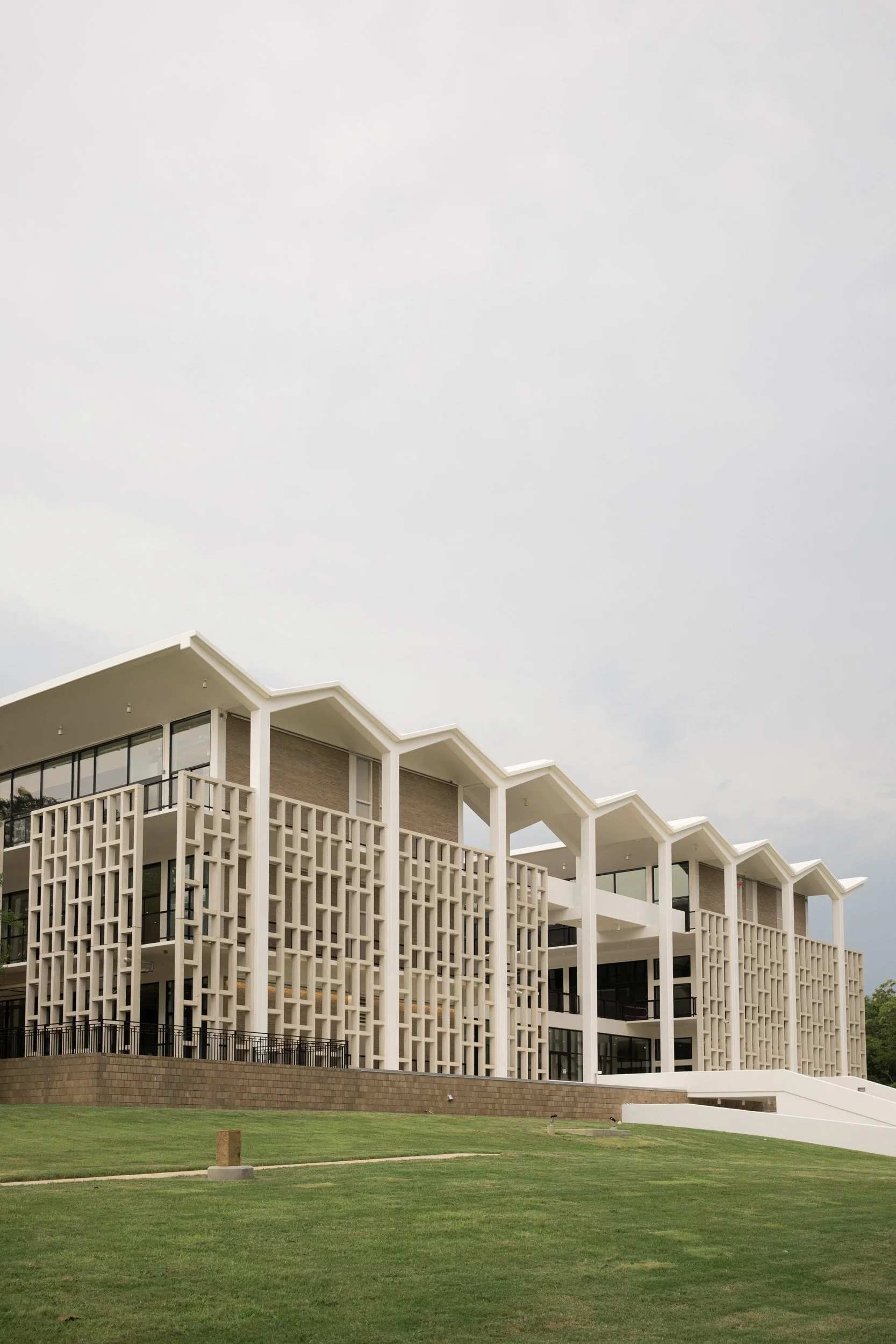 Modern multi-story building with white architectural elements and large windows, set against a cloudy sky with a green lawn in the foreground.