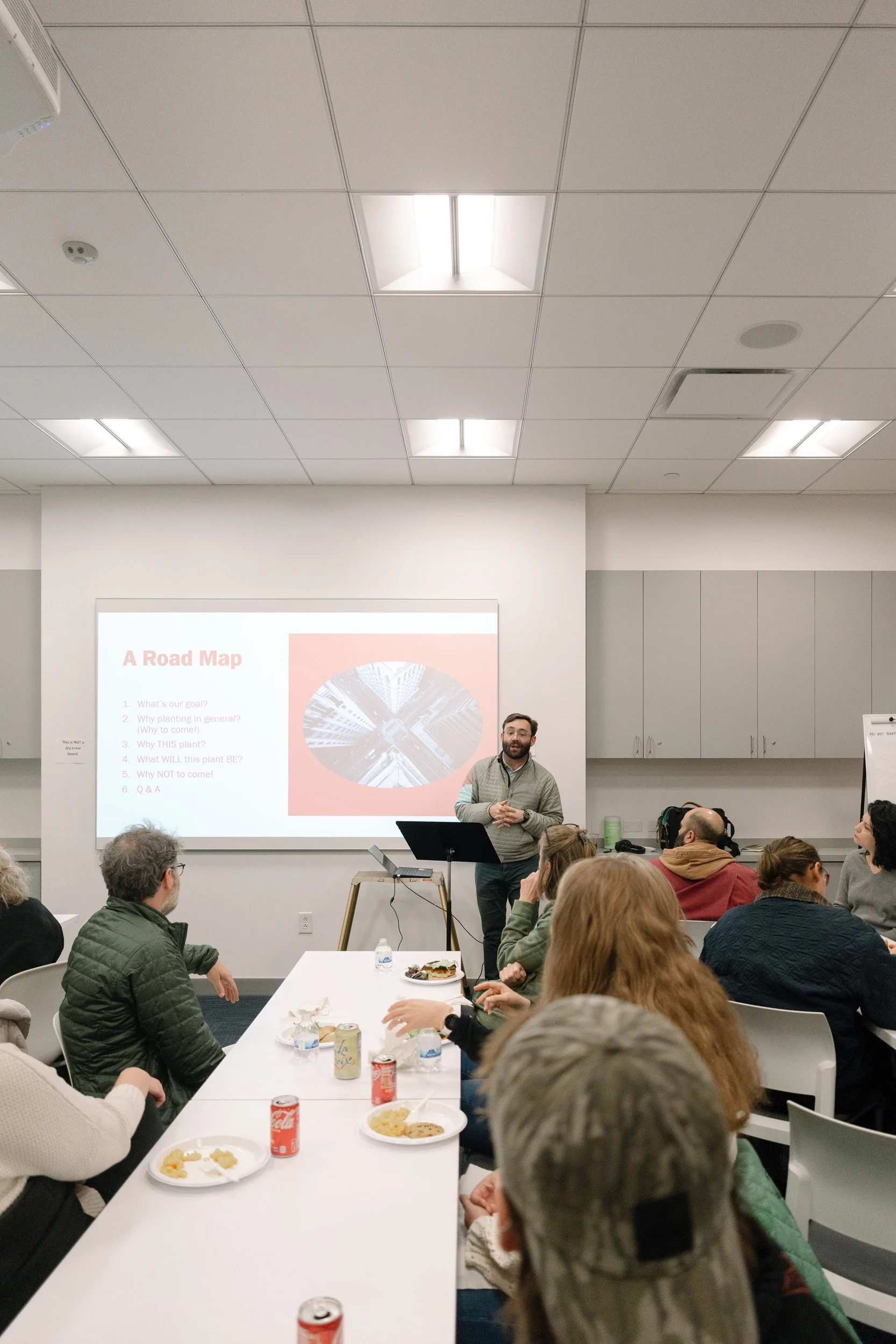 A man with glasses and a beard giving a presentation in a conference room, with an audience of people sitting at tables, some with food and drinks, listening attentively.